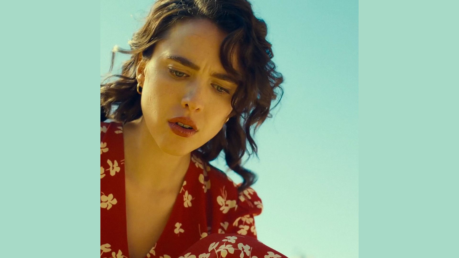Woman in a red floral dress looks down with a thoughtful expression under a clear blue sky. Her curly hair frames her face, suggesting introspection.