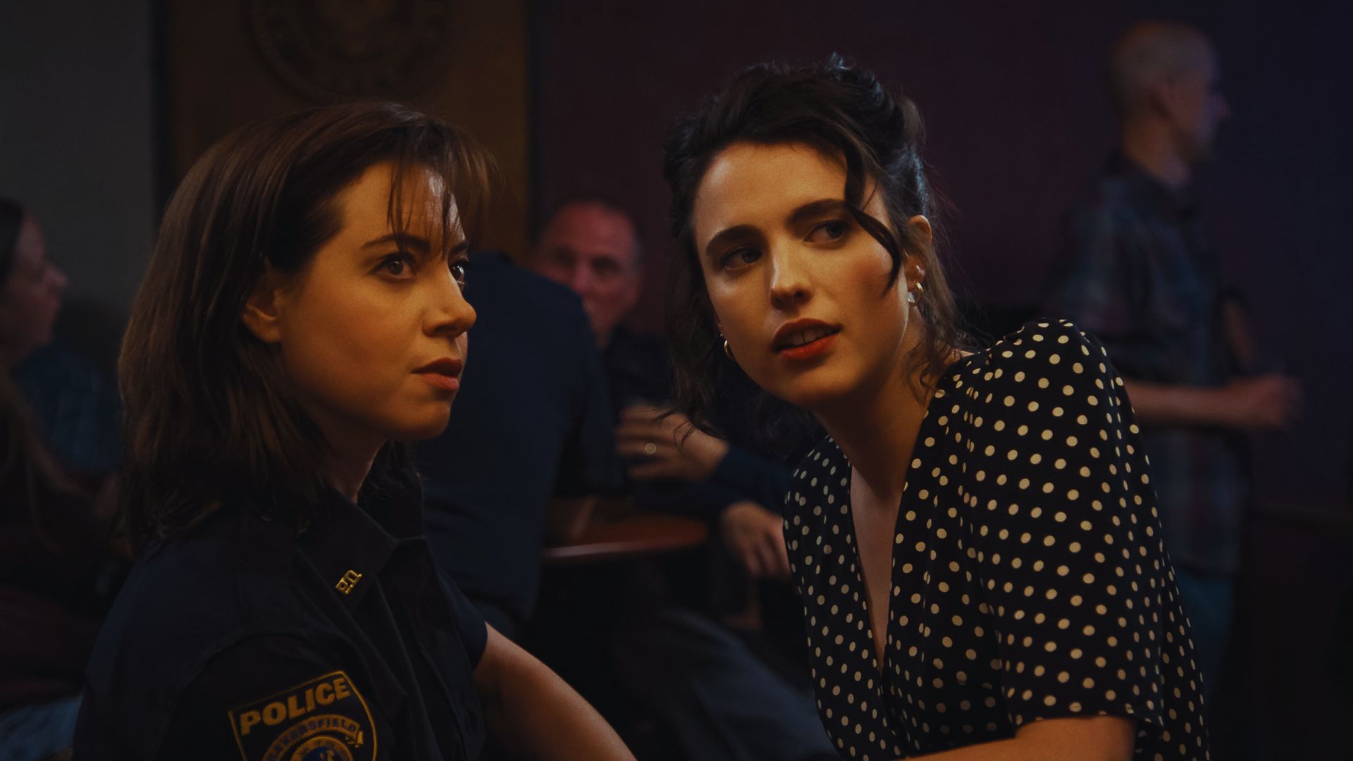 Two women sit in a dimly lit bar, one in a police uniform and the other in a polka dot dress. They appear engaged and focused, creating a tense mood.