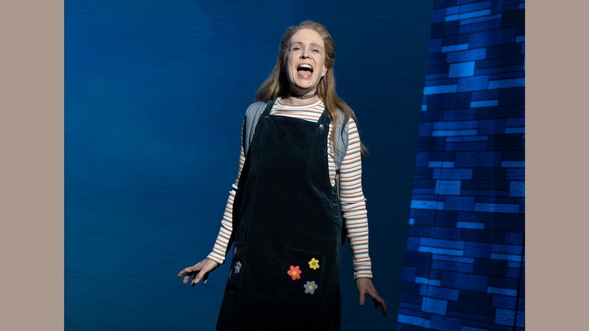 A person on stage sings passionately, wearing a striped shirt and dark overalls with flower patches, set against a blue patterned background.
