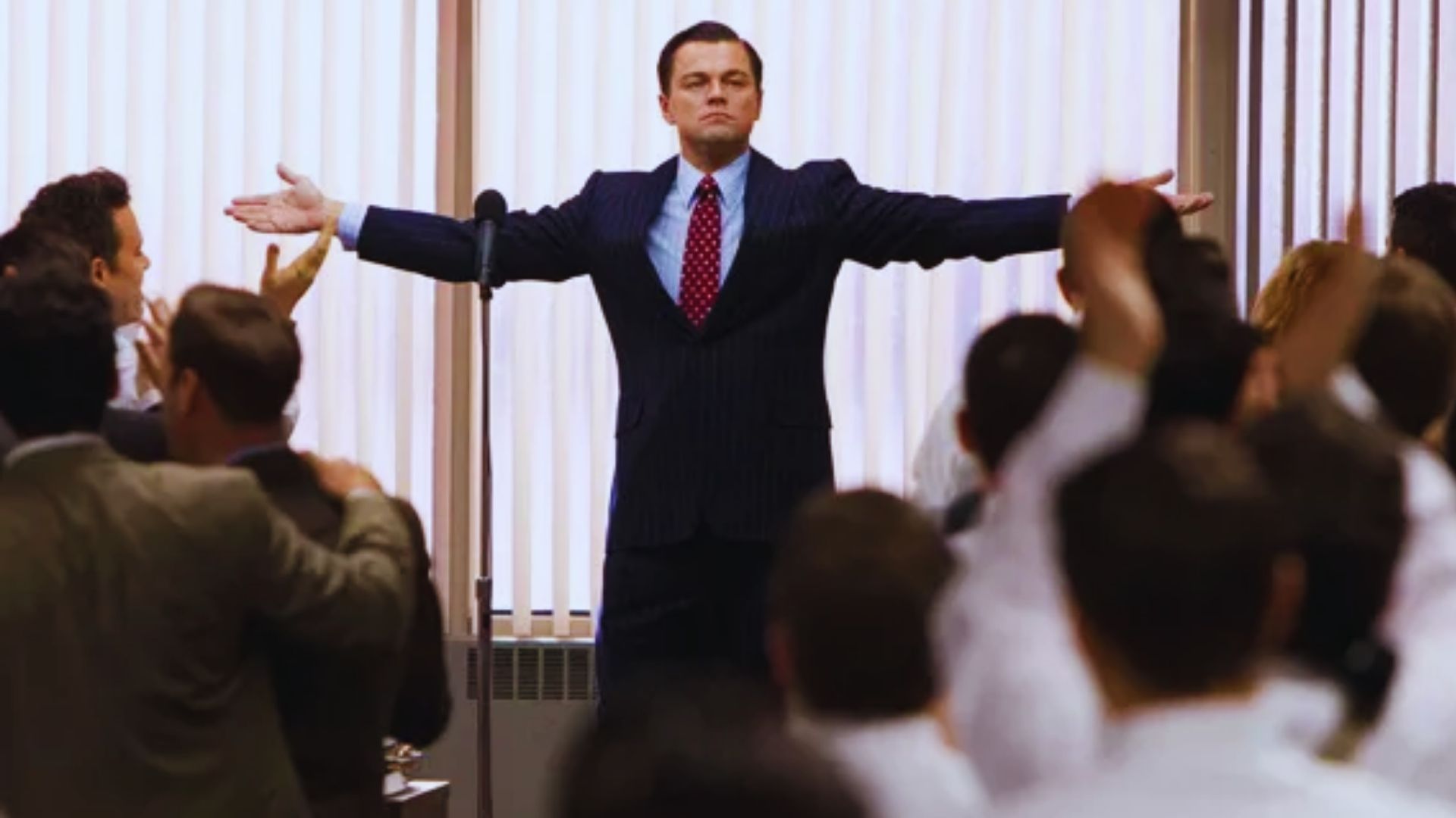 A man in a suit stands confidently with arms outstretched in front of a cheering crowd. The setting appears to be an office with blinds behind him.