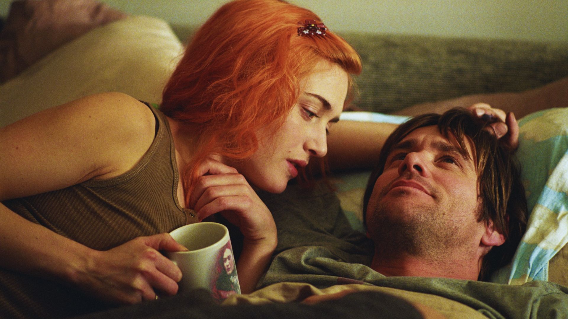 A woman with vibrant orange hair and a tank top leans in intimately towards a man lying down, holding a mug. Both appear relaxed and content.