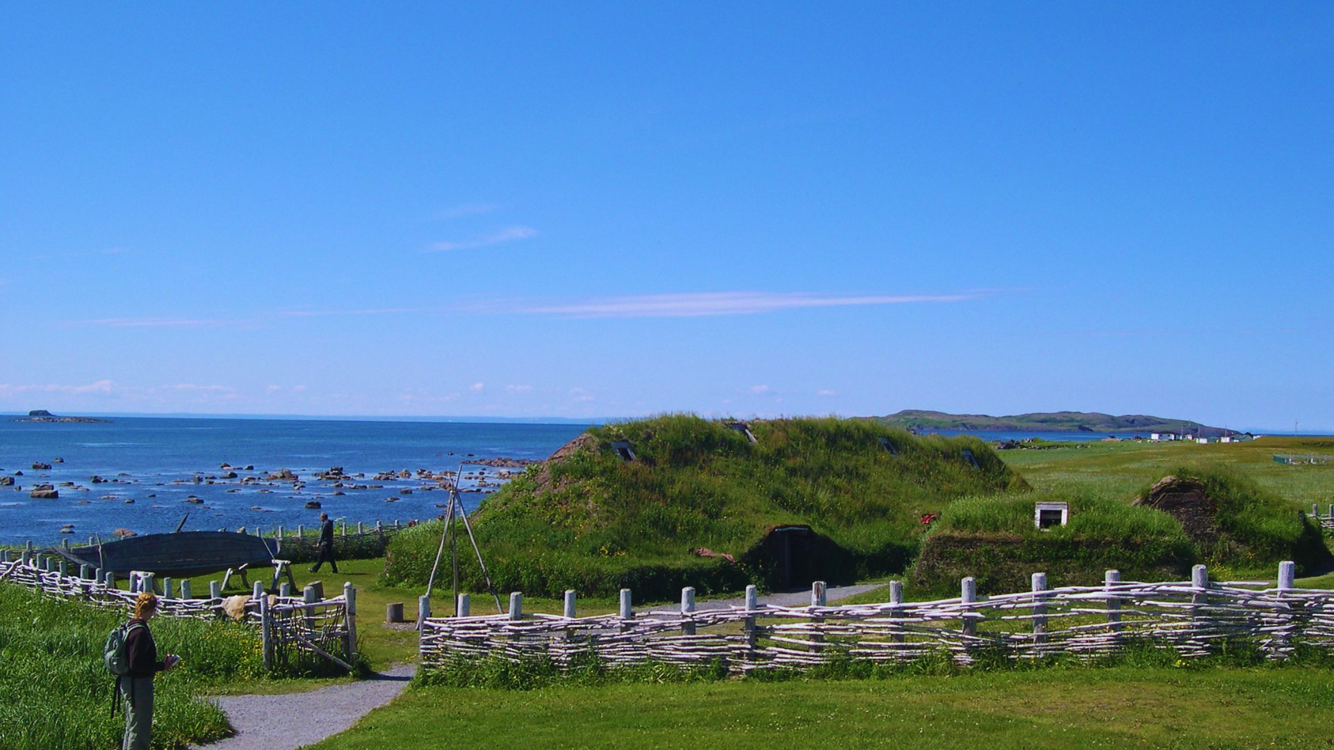 Coastal landscape with grassy hills and turf structures under a clear blue sky. A woven wooden fence surrounds the area, with the ocean in the background.