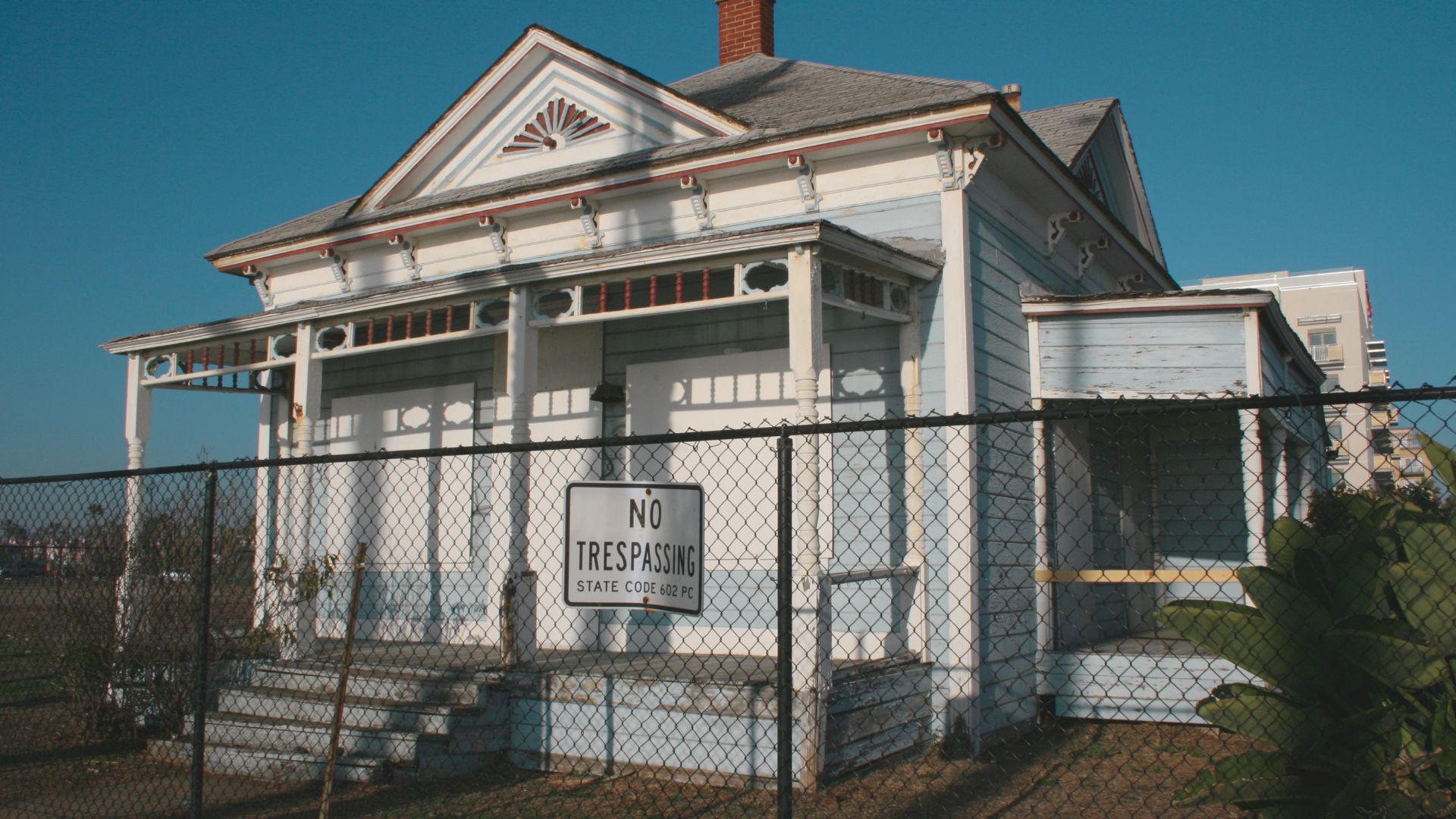 An old white house with a porch and boarded windows is fenced off with a "No Trespassing" sign, conveying a sense of abandonment under a clear blue sky.
