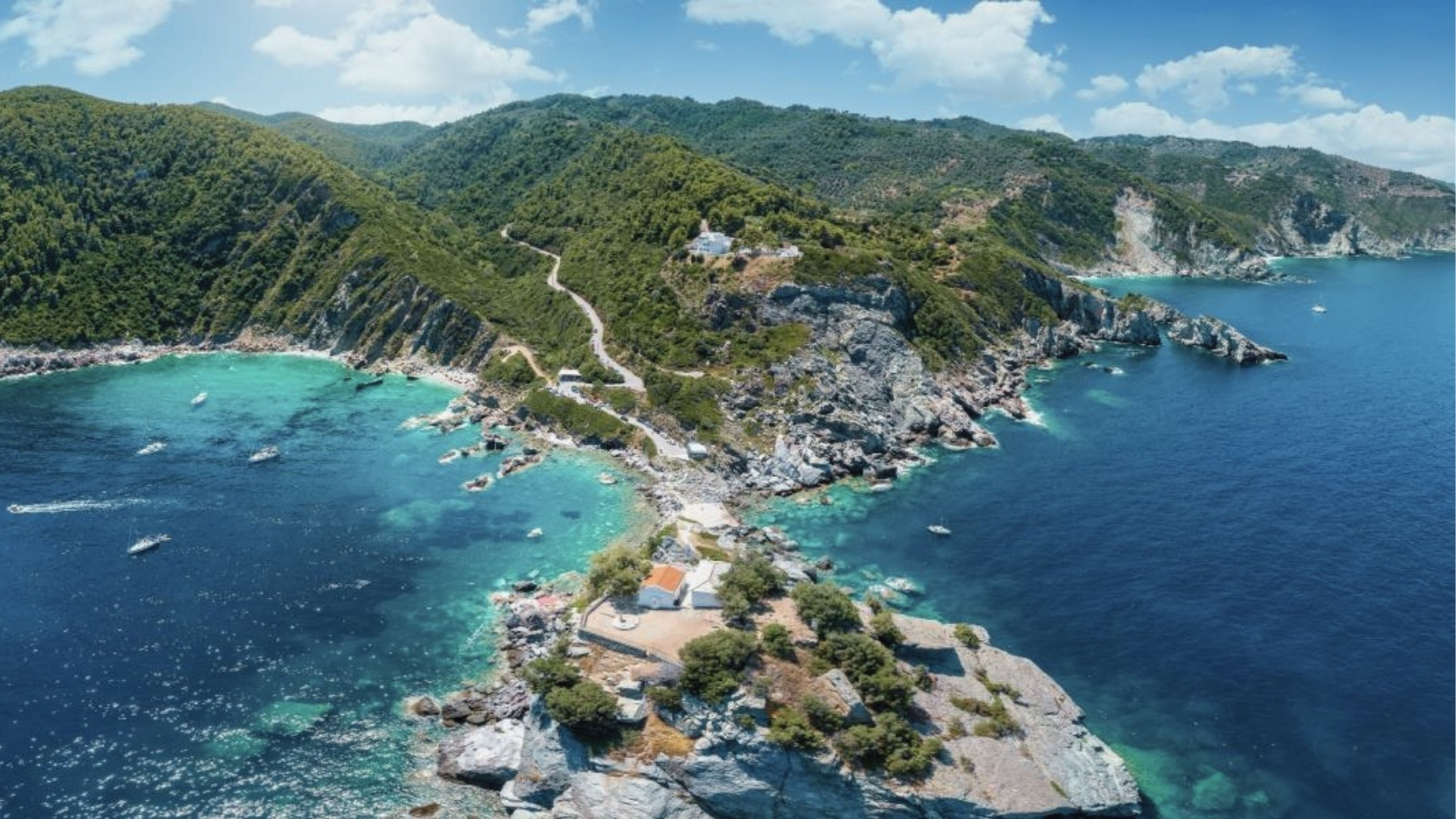 Aerial view of rocky peninsula, turquoise water, boats, winding road, white buildings, green vegetation, and sunny sky in Greece.

