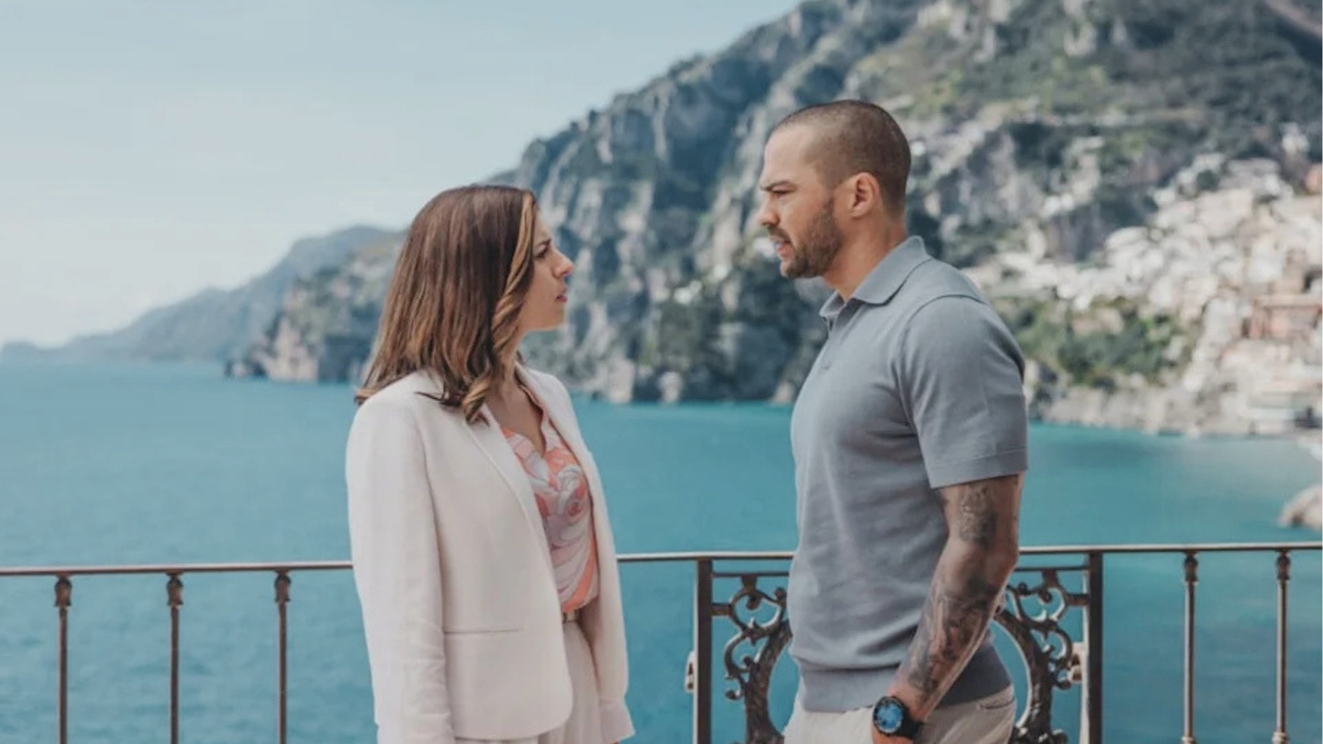 Man and woman talking on balcony overlooking blue sea, dramatic cliffs, Italian coast, Hotel Costiera filming.