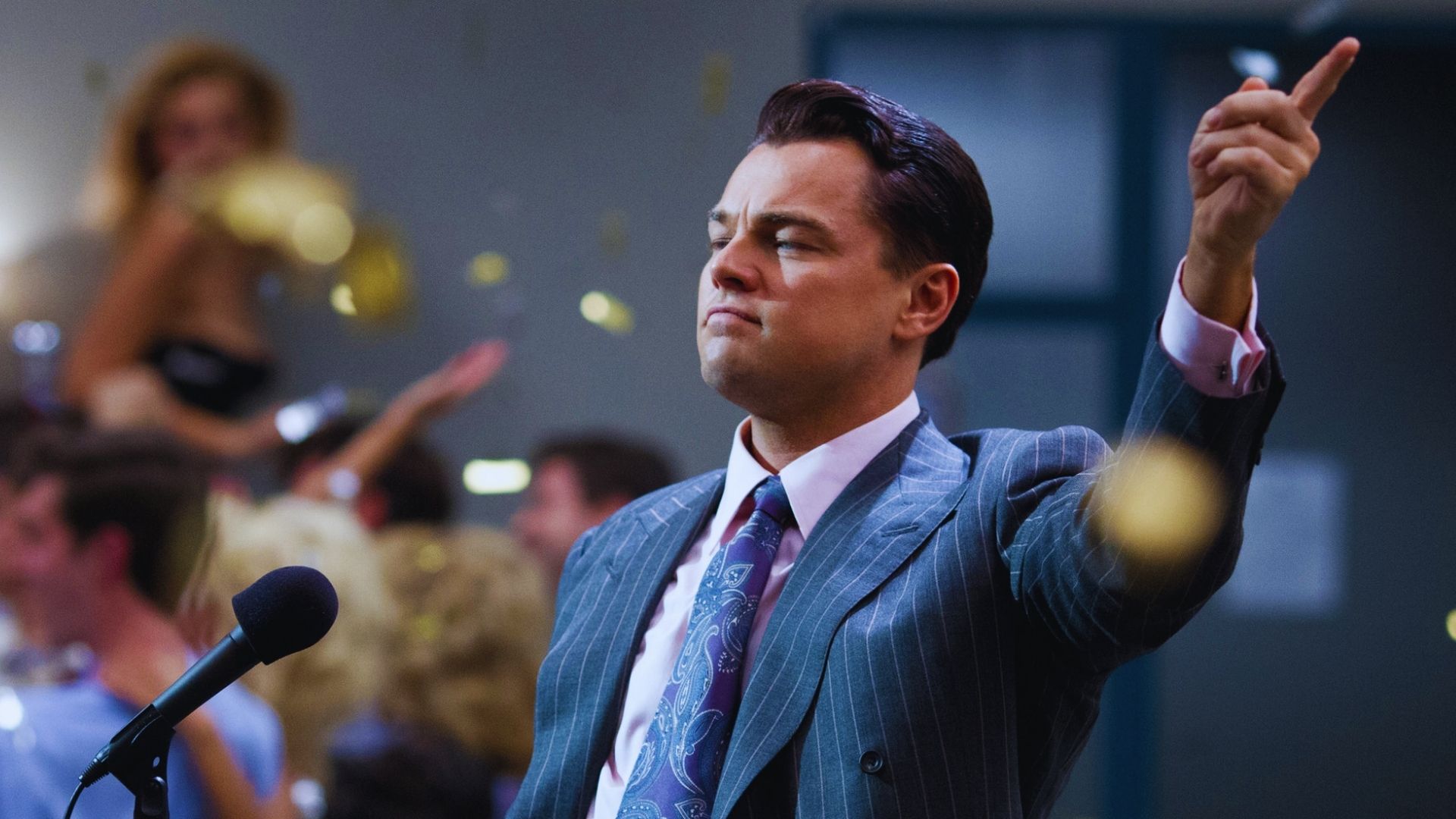 A confident man in a pinstripe suit points upwards while holding a microphone. Gold confetti falls around him, suggesting a celebratory atmosphere.