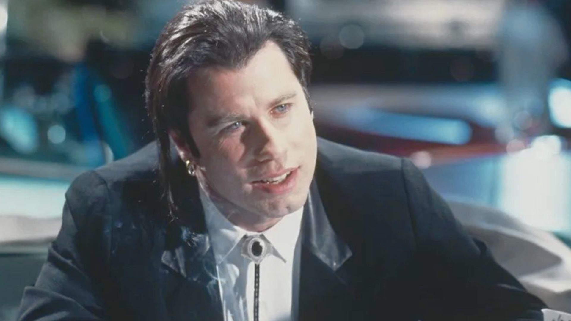 A man in a black suit with slicked-back hair and a bolo tie, sitting in a dimly lit diner. He appears calm and focused, with a slight smile.