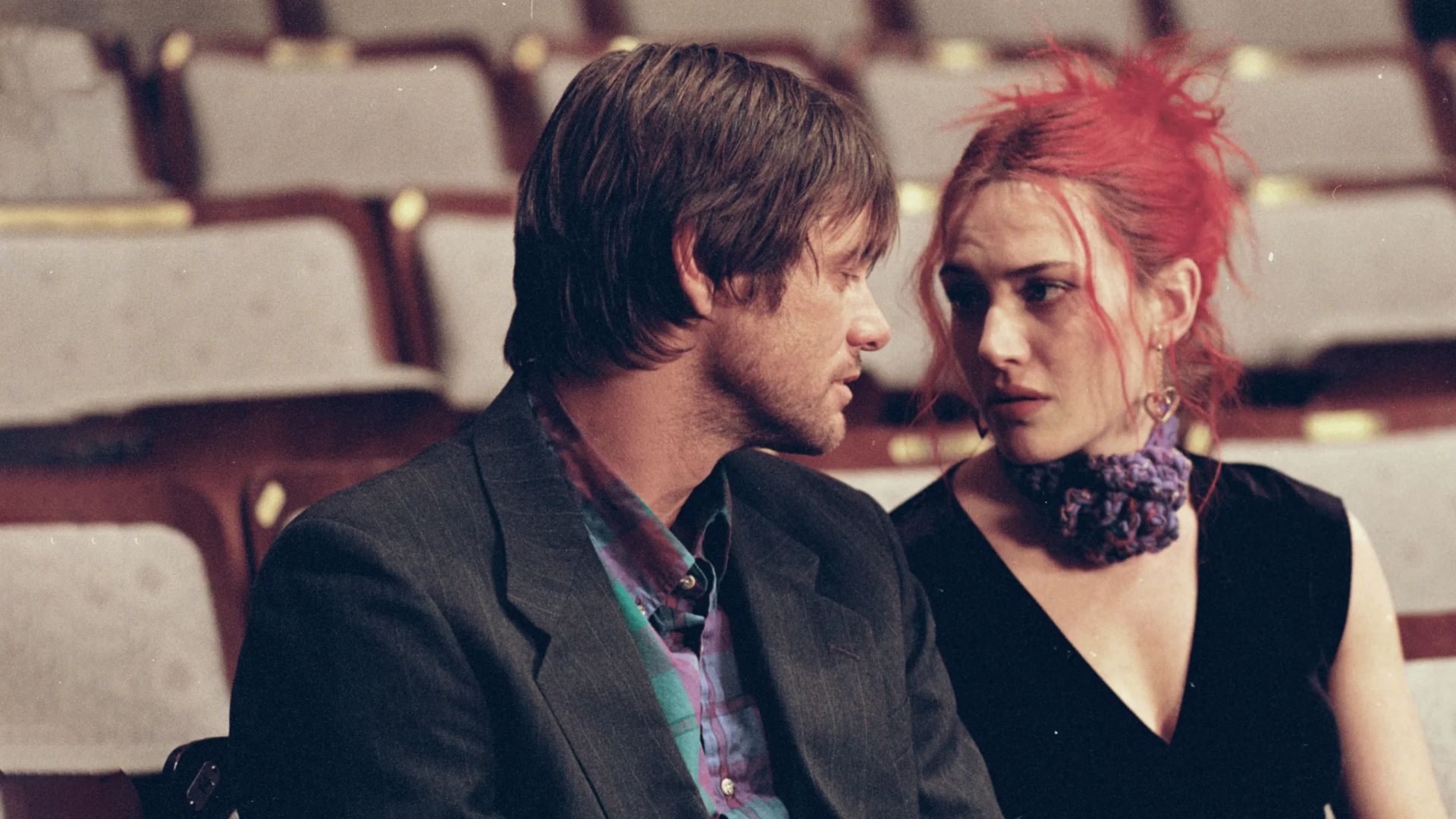 A man in a suit and a woman with red hair sit closely together in a dimly lit, empty auditorium, exchanging an intense, emotional gaze.