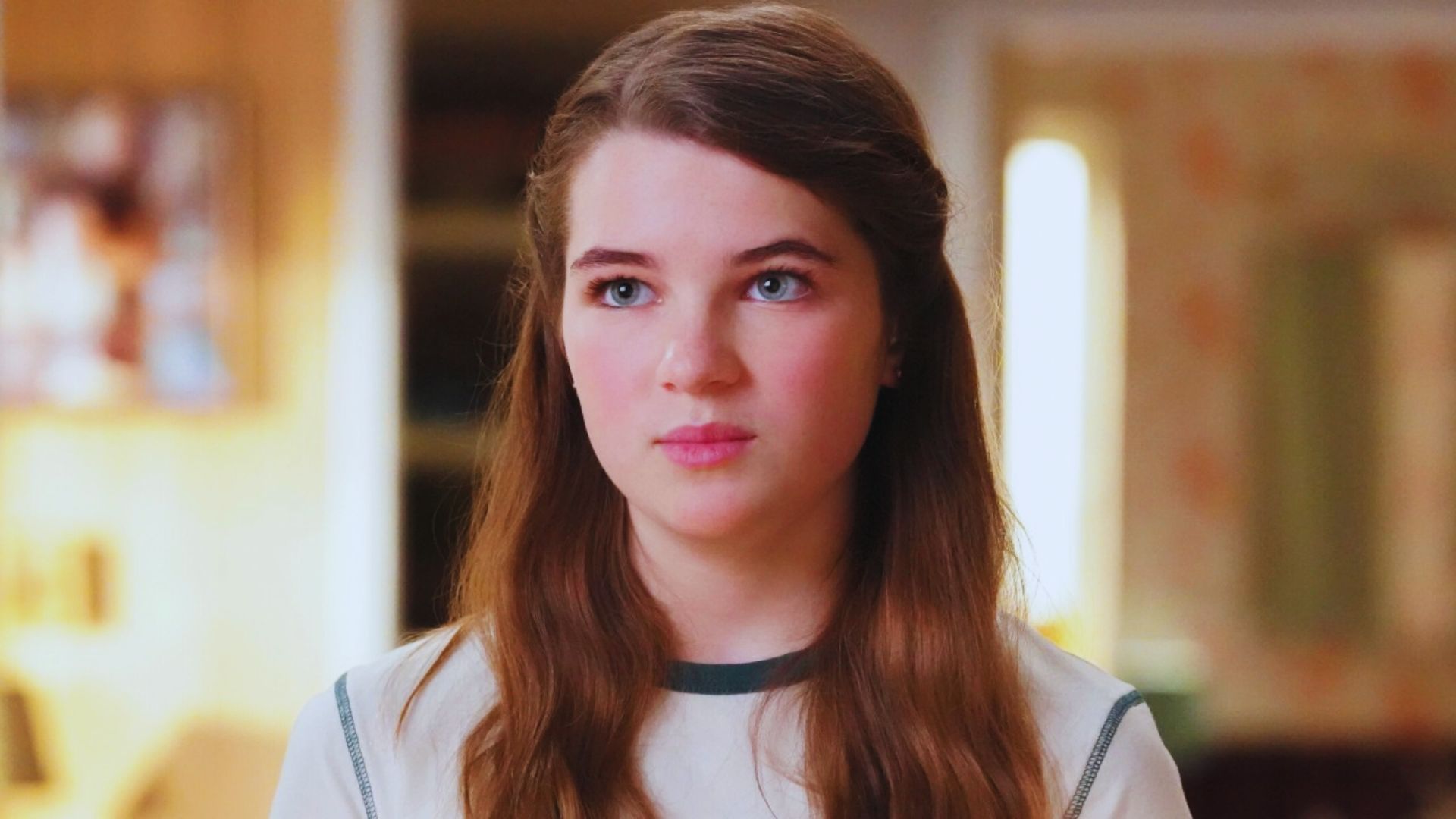 A young woman with long brown hair and a thoughtful expression stands in a softly lit room with blurred, warm-toned decor in the background.