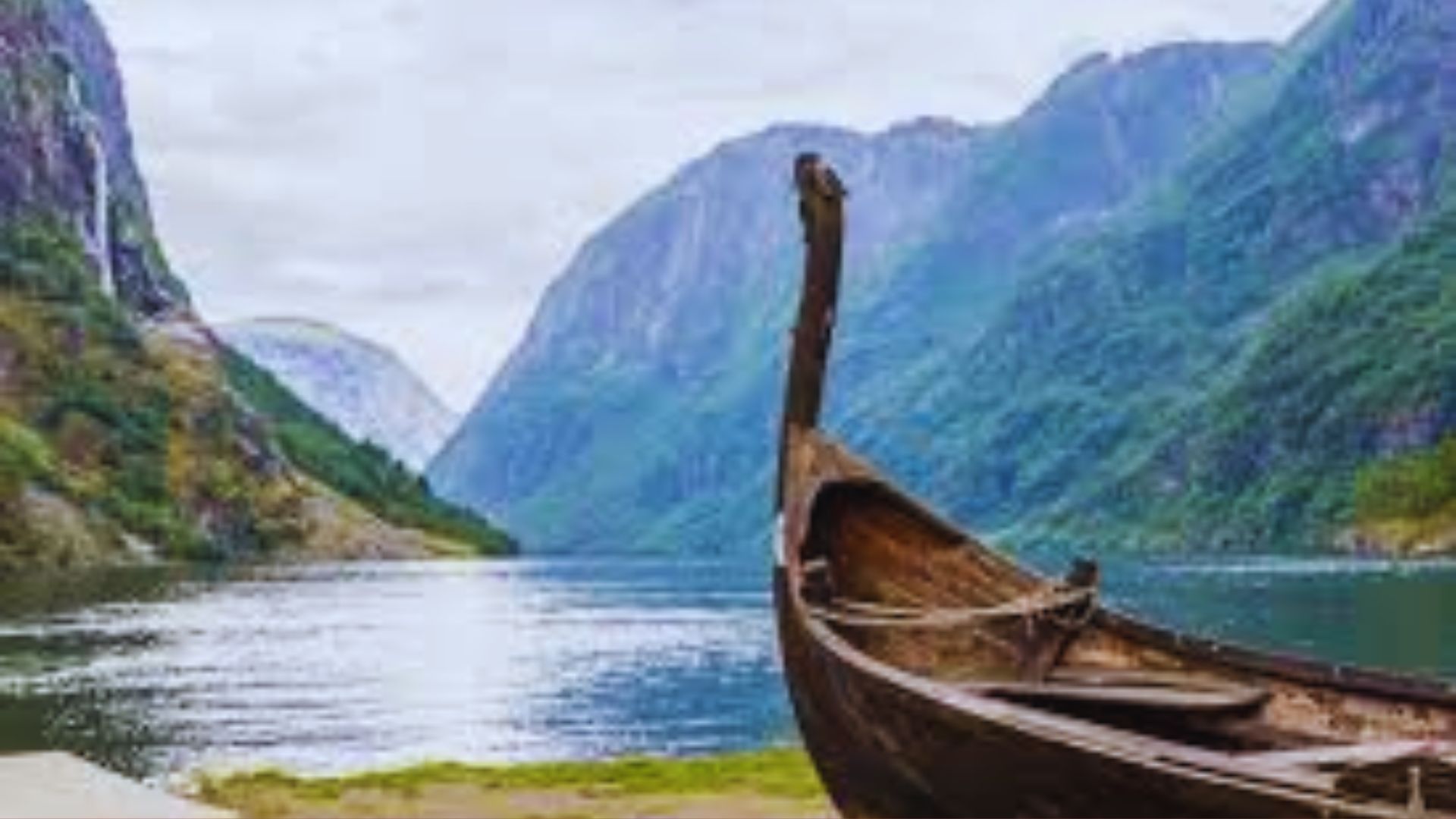 A wooden Viking ship rests on the shore of a serene fjord, surrounded by steep, green mountains under a cloudy sky. The scene is peaceful and majestic.