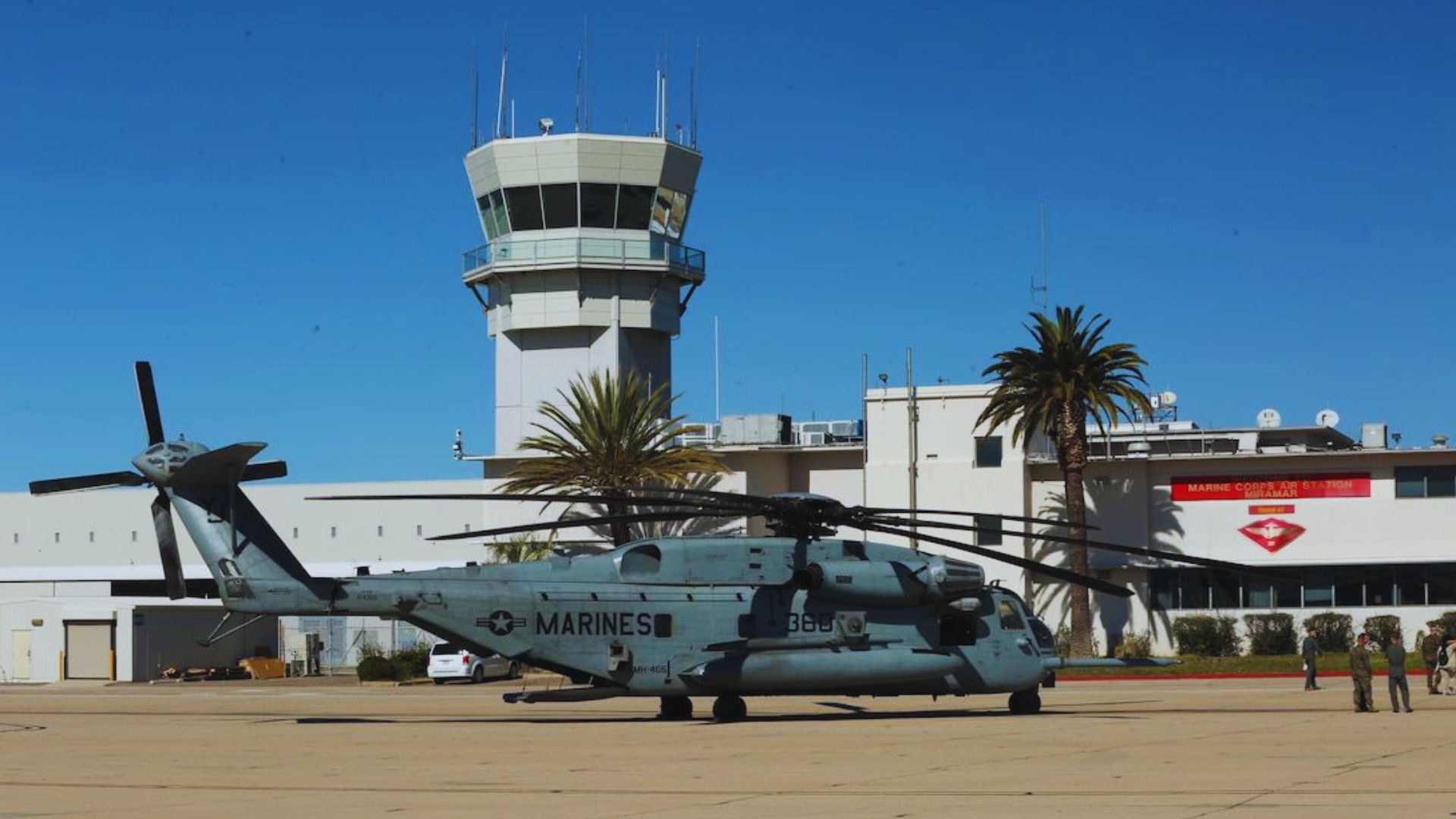 A military helicopter labeled "MARINES" is parked on a runway with tall control tower and buildings in the background. Palm trees frame the scene under clear blue skies.