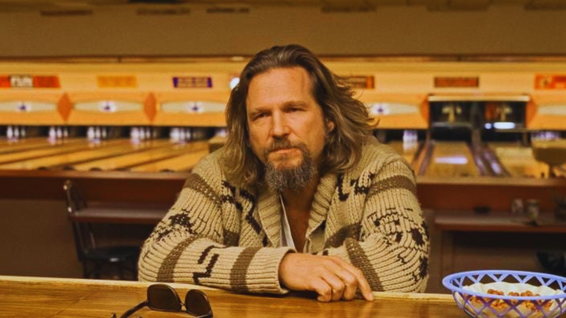 A man with long hair and a beard leans on a wooden counter in a bowling alley, wearing a patterned cardigan. Sunglasses lie nearby, evoking a relaxed mood.