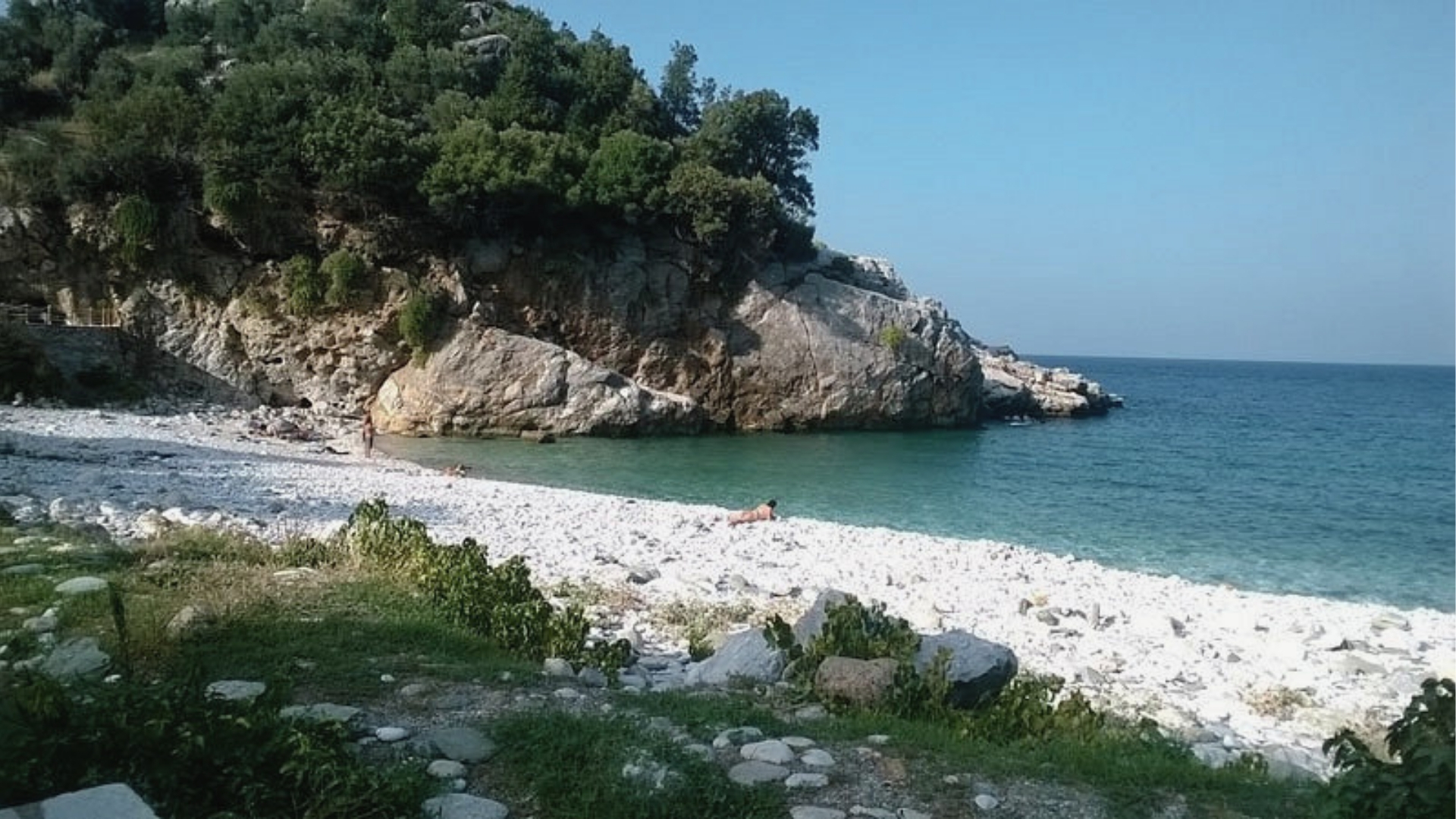 Rocky cliff covered with dense vegetation, white pebble beach, turquoise water, bright sky, and people relaxing in Greece.
