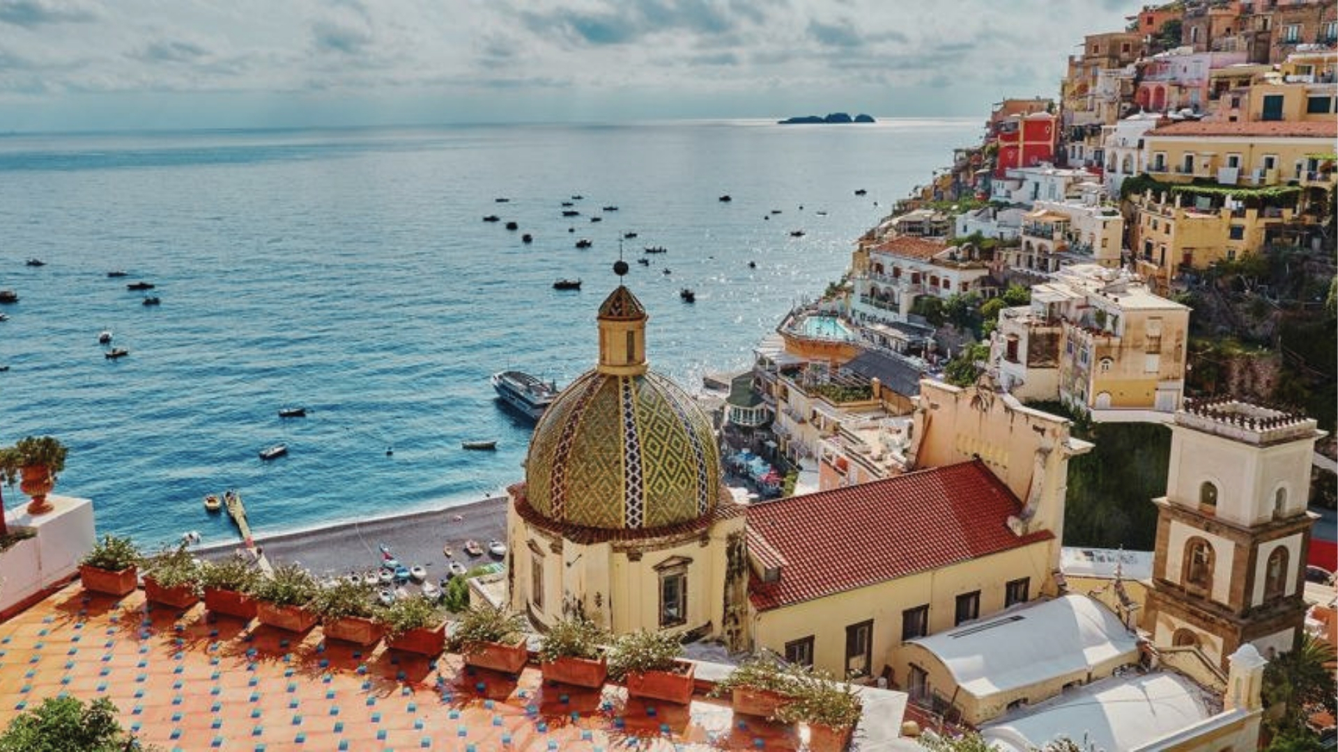 Positano church with tiled dome, black sand beach, colorful buildings, boats, blue sea, Hotel Costiera filming location.
