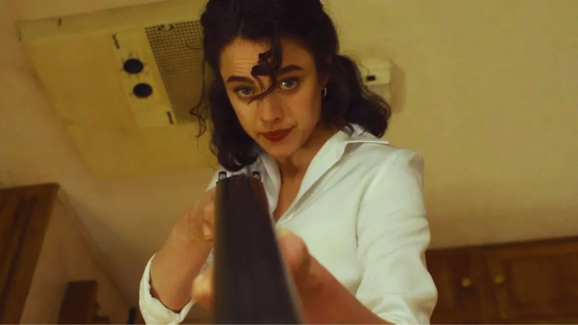 Woman in a white shirt points a gun downward with a focused expression. The image has a tense and dramatic tone, set in a wooden interior space.