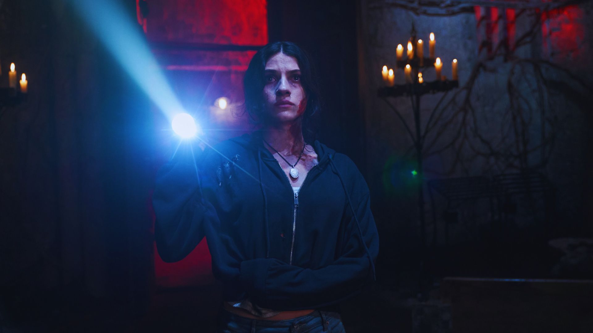 A woman stands in a dimly lit room, holding a flashlight that beams brightly. She appears tense. Lit candles in holders cast a warm glow on the stone walls.