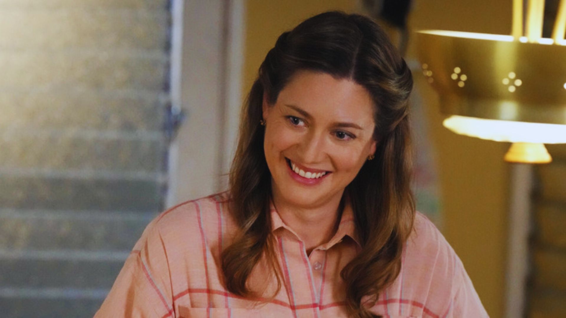 Smiling woman with wavy brown hair in a pink plaid shirt, stands in warm ambient light, suggesting a cozy and cheerful atmosphere.