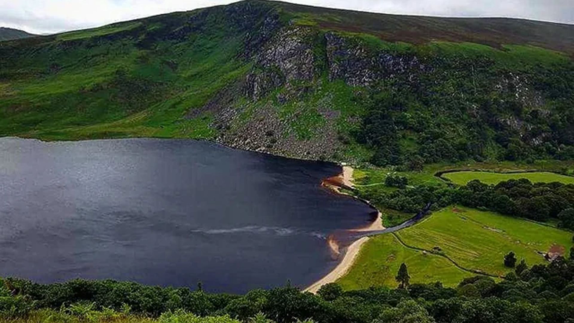 A serene lake bordered by lush green hills under a cloudy sky. A small sandy beach curves around one edge, contrasting with the dark water. Peaceful and scenic.