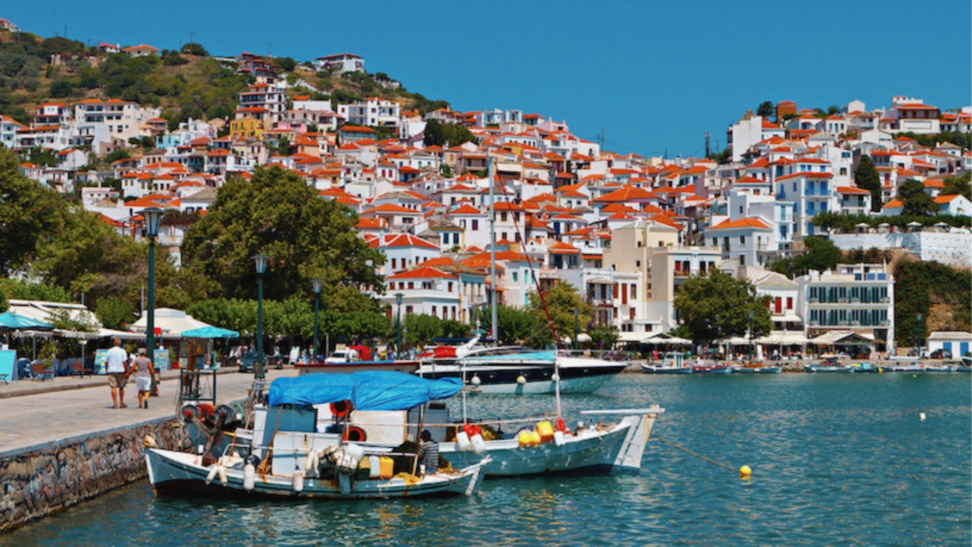 Coastal town hillside with white buildings, red-tiled roofs, harbor boats, promenade, umbrellas, lampposts, and clear blue sky in Greece.