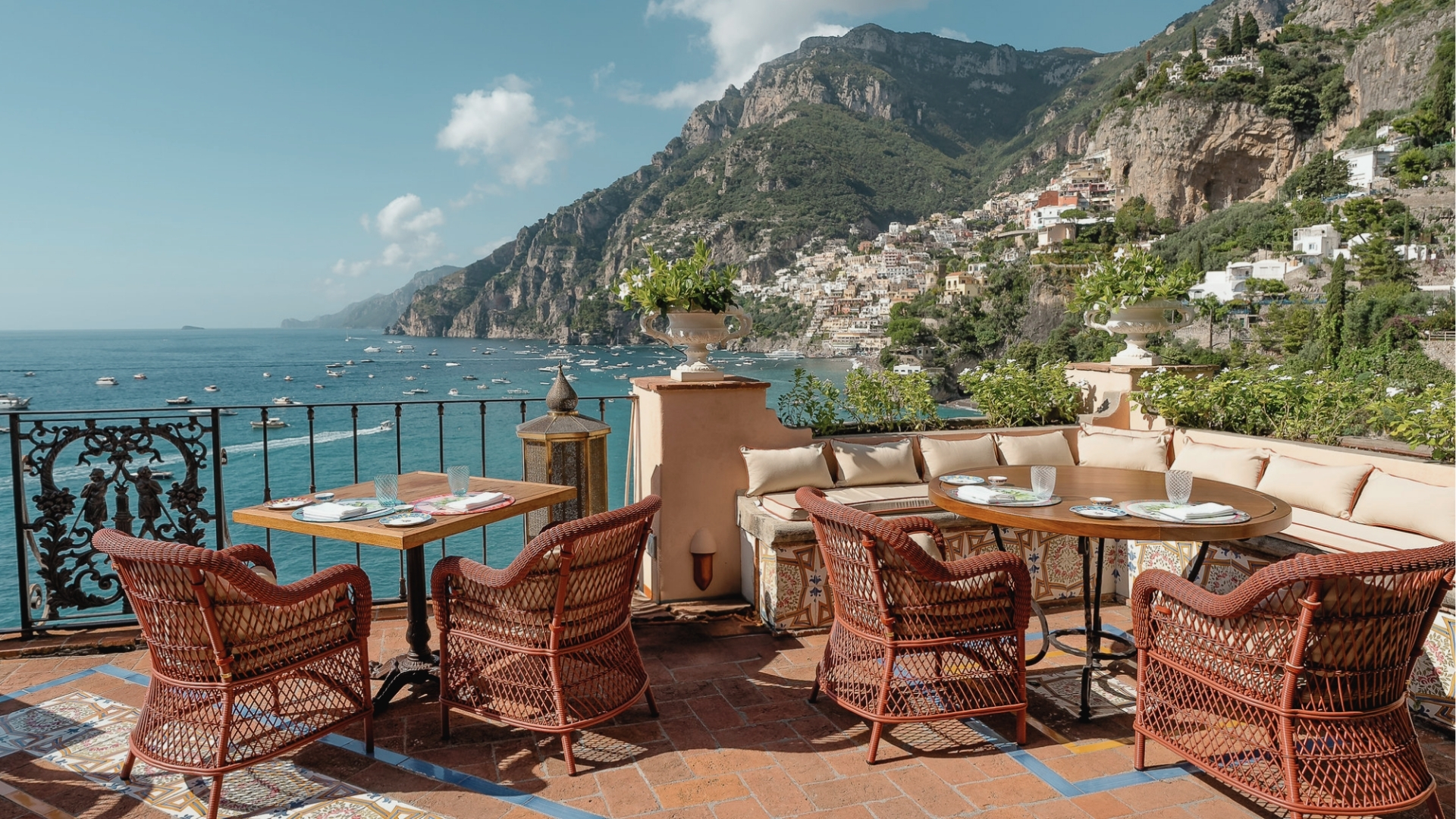 Seaside terrace with wicker chairs, set tables, overlooking boats, mountains, cliffside village, Hotel Costiera filming location.
