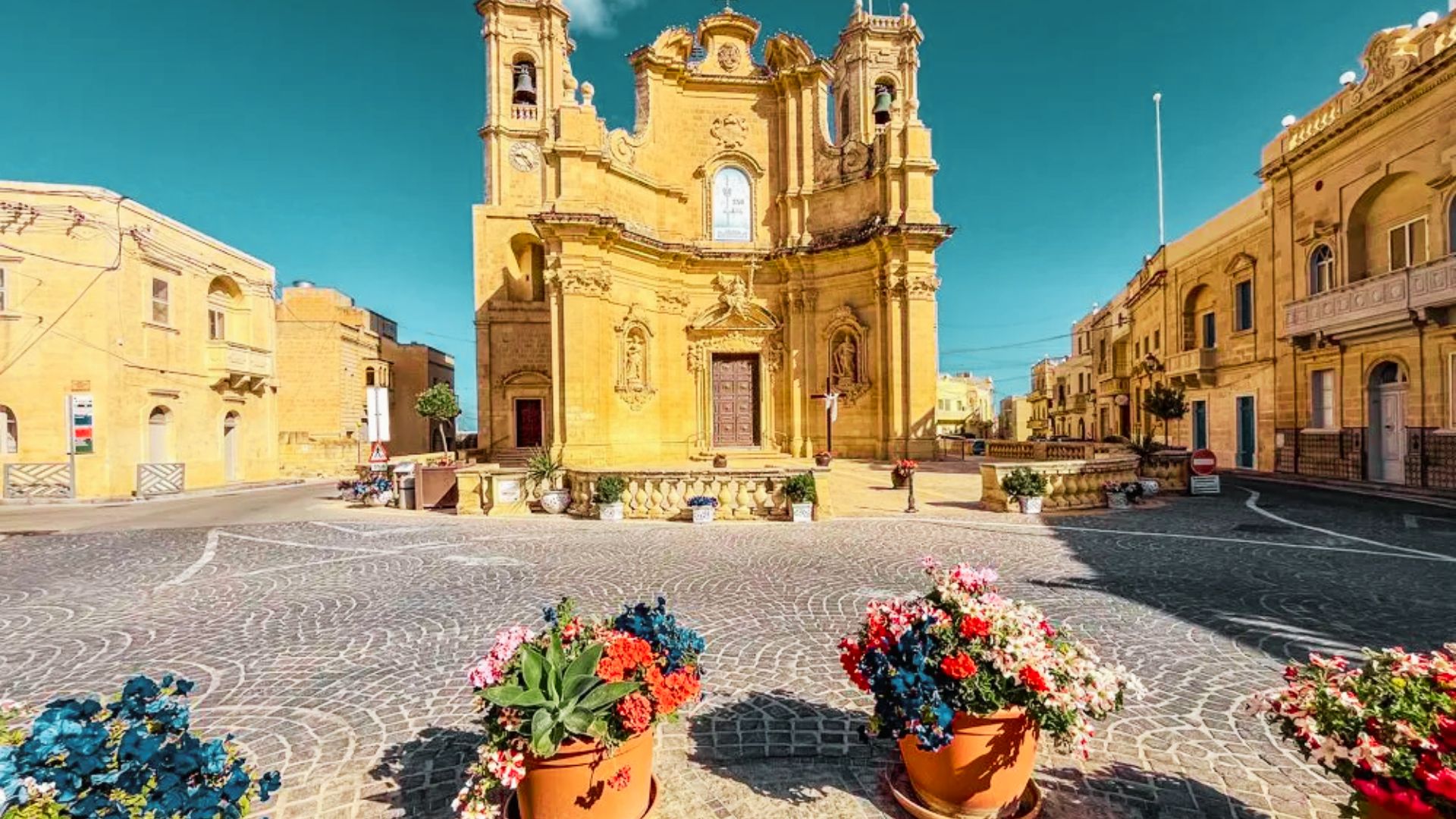 A church surrounded by flower pots on the ground, featured in the Madame Blanc filming locations in Gozo.
