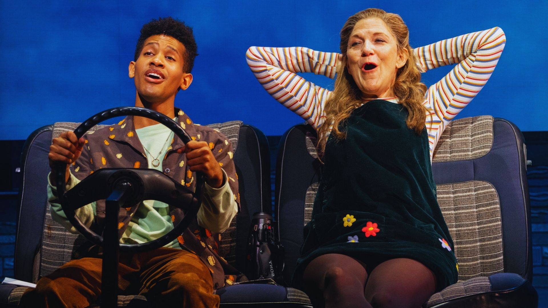 A young man energetically pretends to drive, holding a steering wheel, while an older woman sits beside him, relaxed with hands behind her head, both emoting joy on a staged setting.