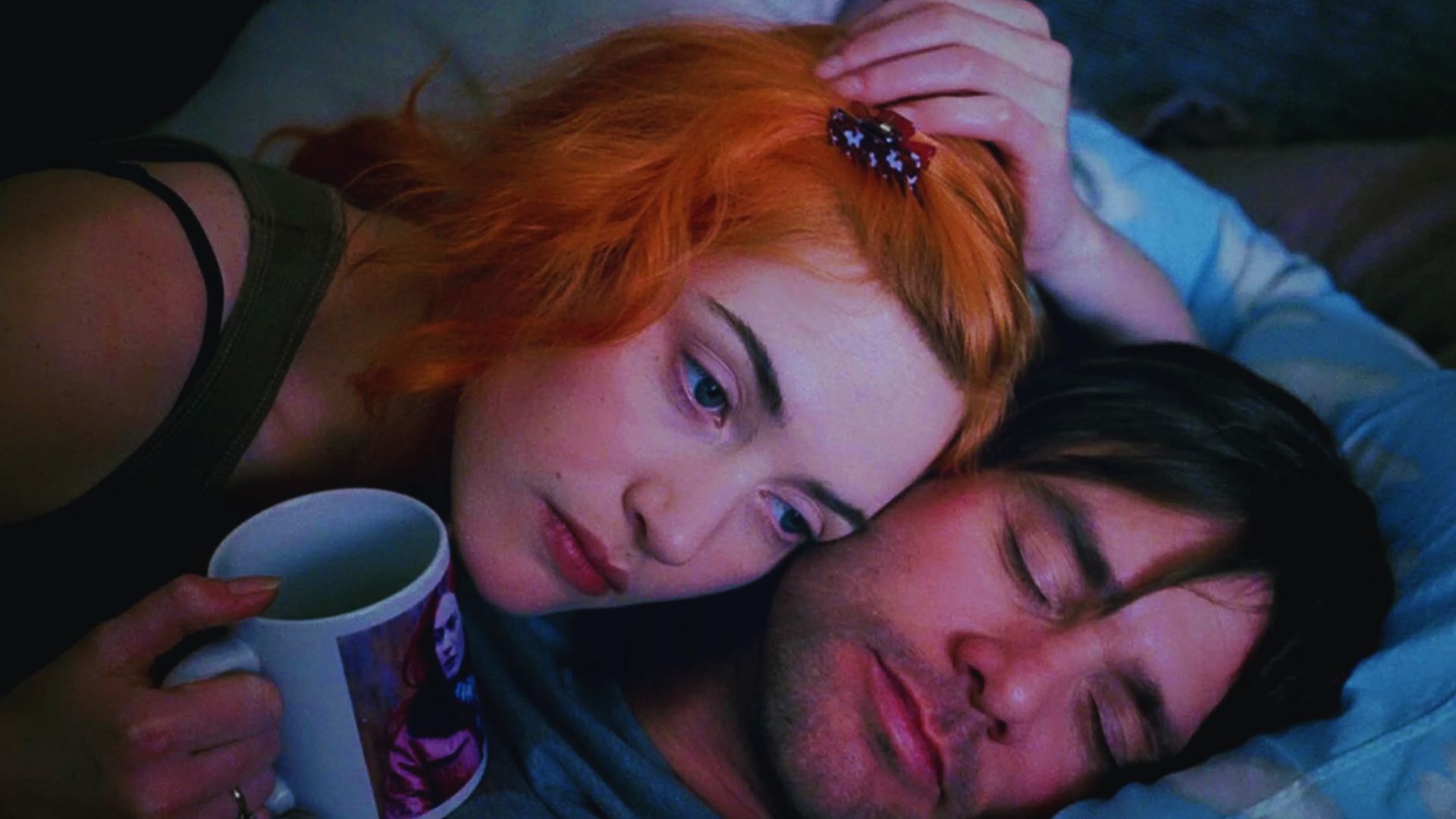 A woman with bright orange hair and a hair clip holds a mug and rests her head gently on a man's shoulder as he sleeps, creating a calm, intimate mood.
