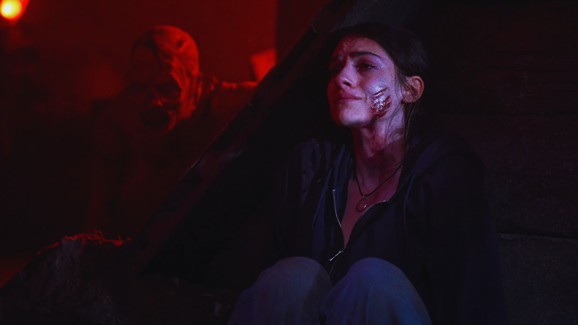A woman with a wounded cheek sits in fear against a dark backdrop, dimly lit in red. A menacing figure lurks behind her, enhancing the tense atmosphere.