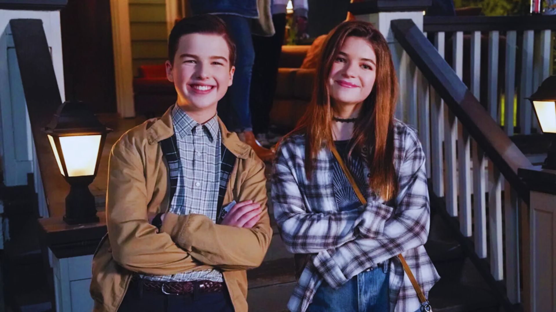 Two teenagers stand on a porch, smiling with arms crossed. Both wear plaid shirts; the boy has a tan jacket. The scene is warmly lit, creating a cheerful vibe.