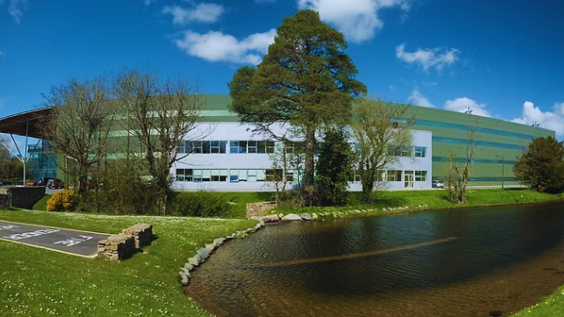 A modern industrial building with green and white stripes stands beside a small pond under a clear blue sky, surrounded by trees and grass.