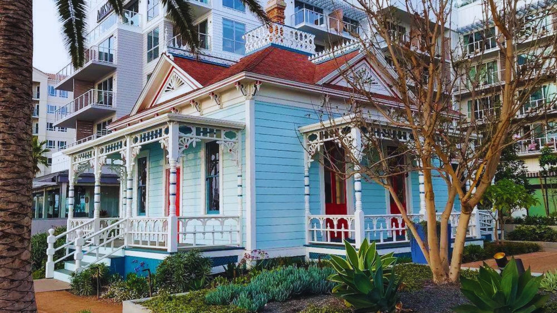 A charming, light blue Victorian house with intricate white trim and a red roof is surrounded by modern buildings, trees, and landscaped greenery.