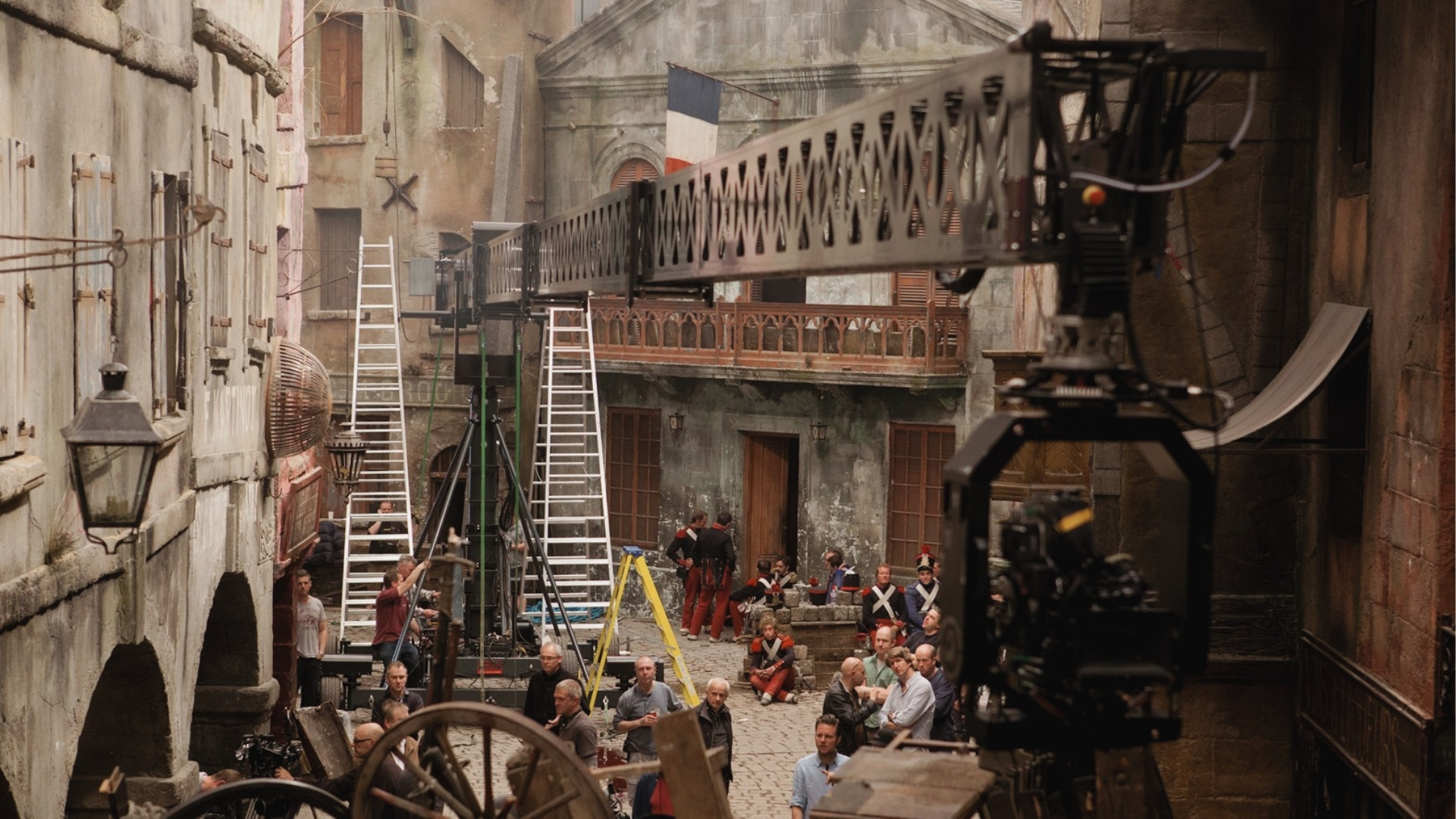 Film set resembling old European street, stone buildings, French flag, camera crane, crew, and actors in London.
