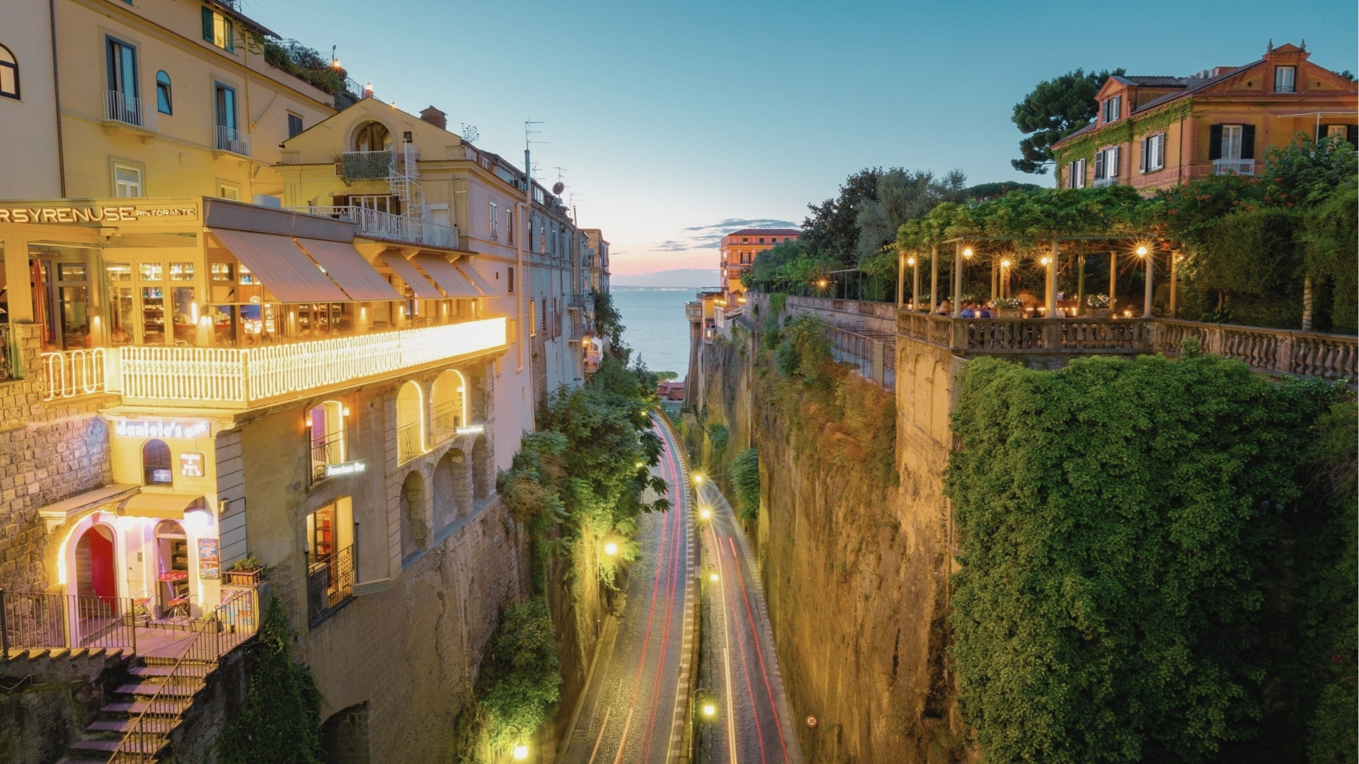 Cliffside restaurants and terraces glow at dusk above winding coastal road, sea, Hotel Costiera filming location.
