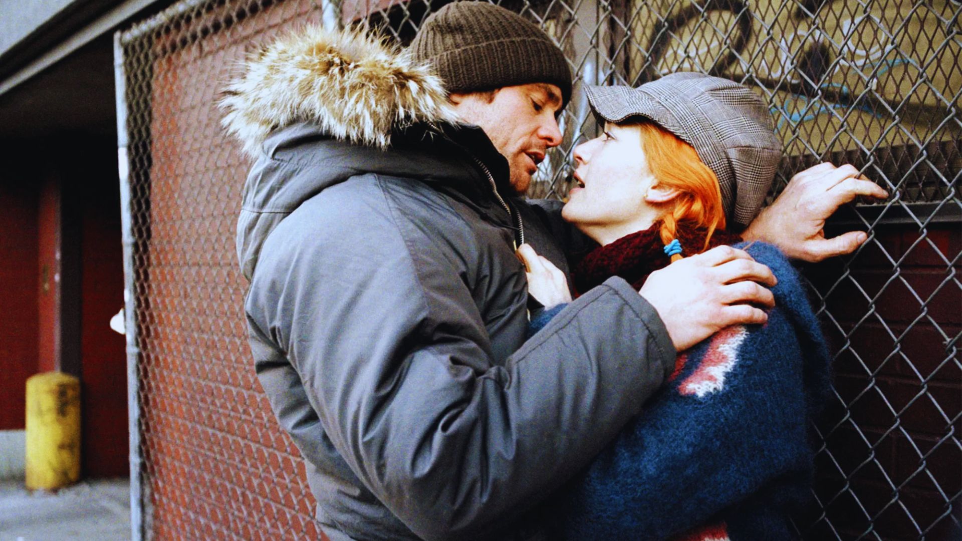 A couple stands closely against a chain-link fence. The man in a winter coat and beanie leans in towards the woman, who has orange hair and a cap. Intimate and emotional.