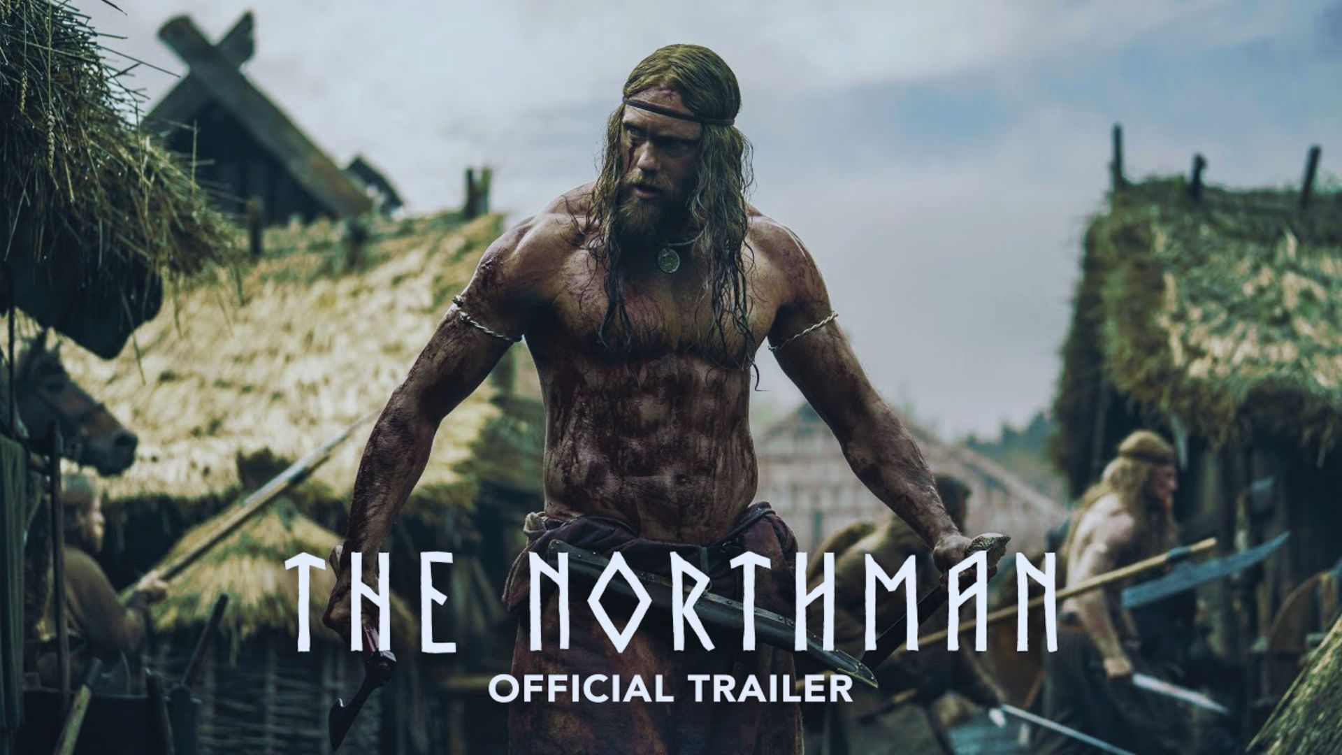 A bloodied warrior with long hair stands fiercely in a Viking village, surrounded by thatched huts and other warriors, conveying tension and intensity. Text reads "The Northman Official Trailer."