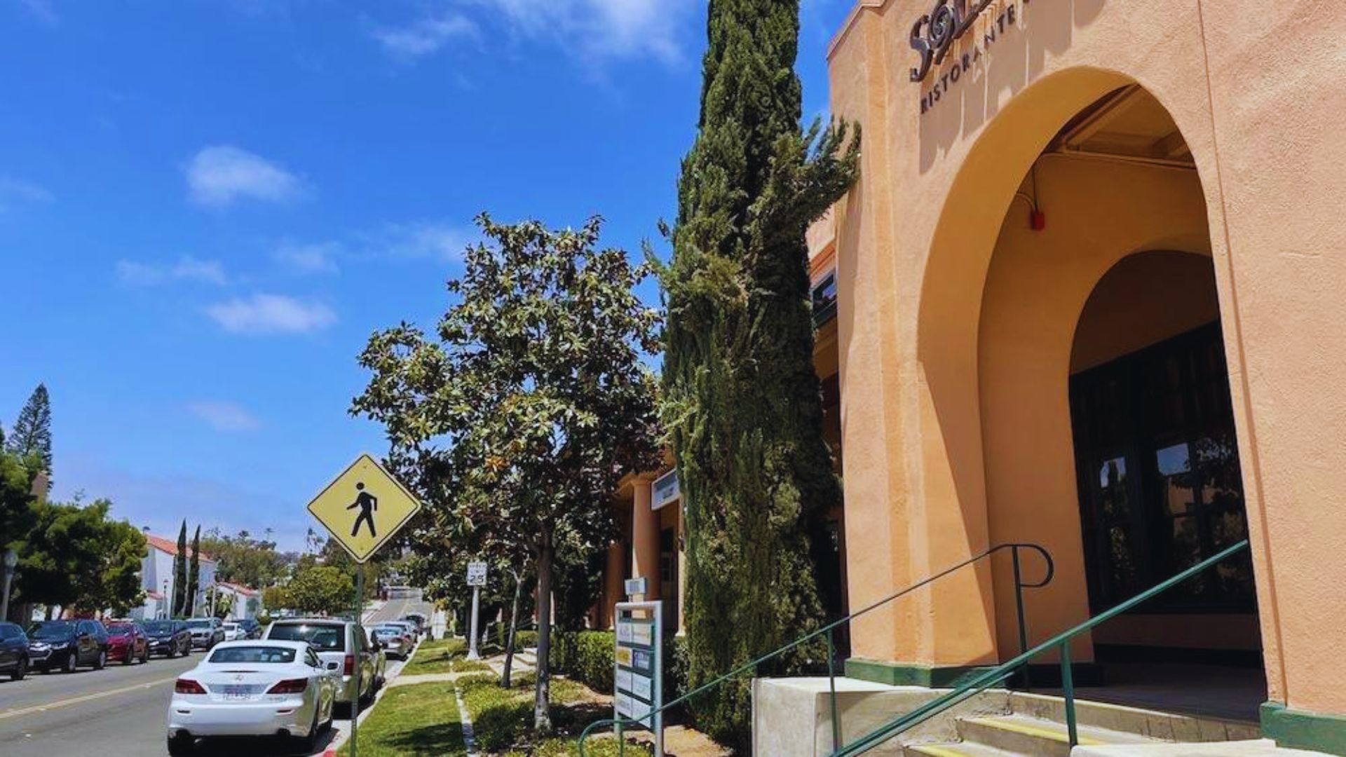 Street scene with a pedestrian crossing sign, parked cars, and a peach-colored building with arches. Trees and a clear blue sky create a relaxed atmosphere.