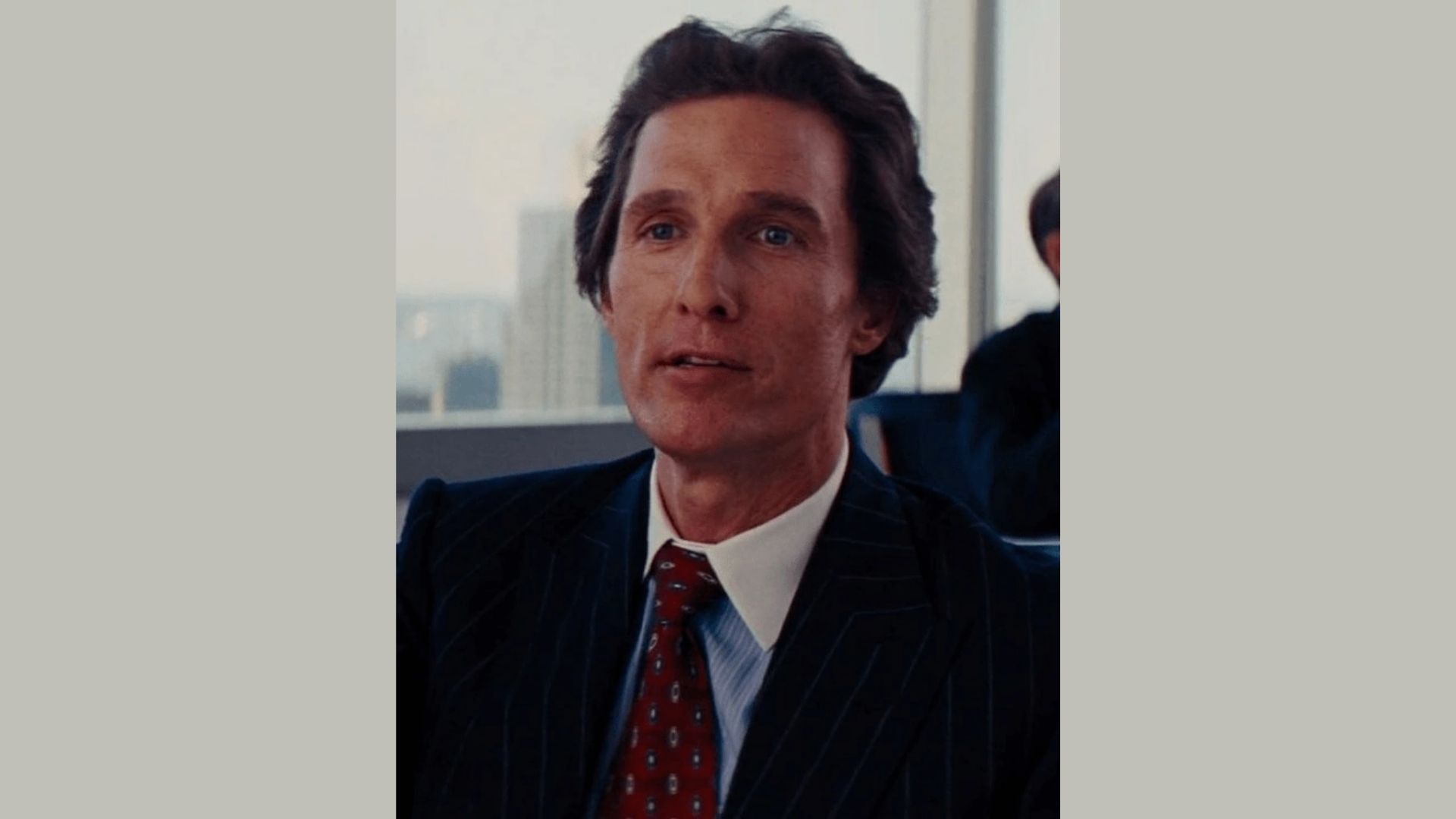 A person wearing a dark pinstripe suit, white shirt, and red tie sits in an office with large windows behind, conveying a professional and serious tone.