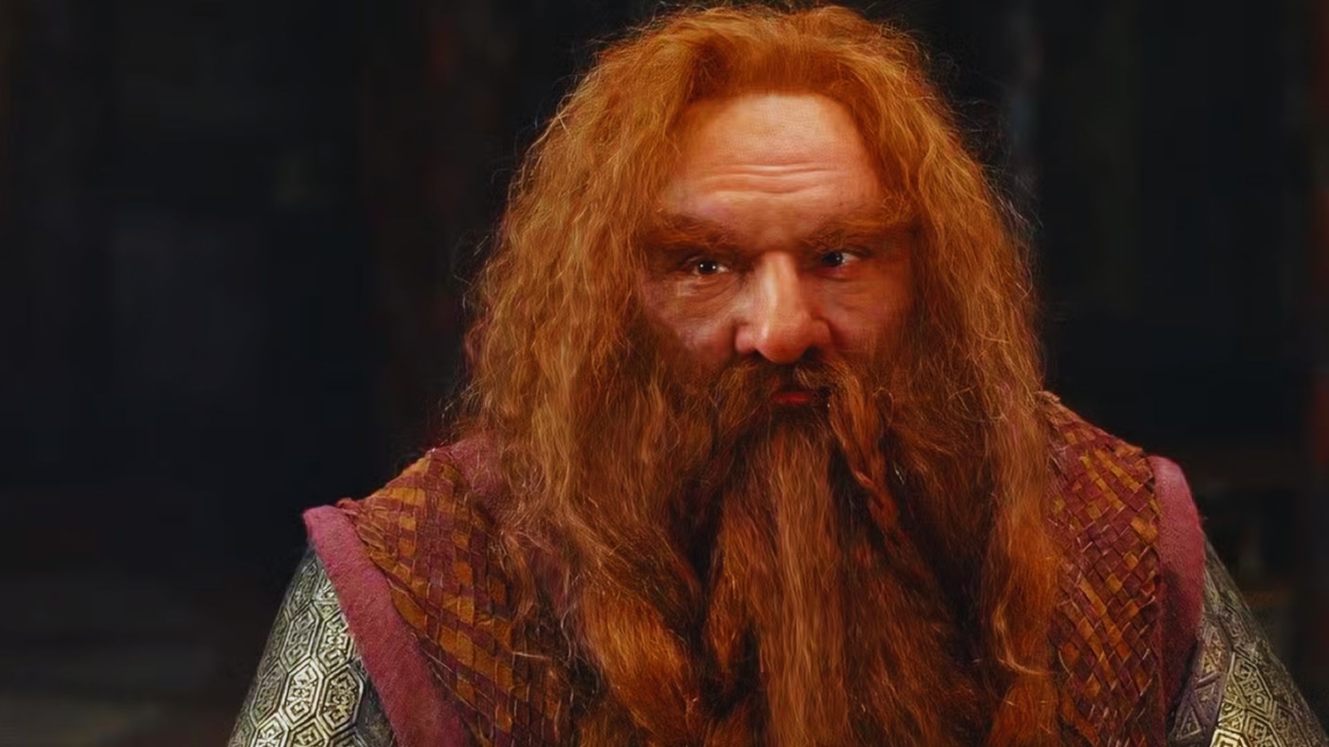 A dwarf with long red hair and a braided beard sits indoors wearing ornate armor and a patterned cloak. His expression is serious and contemplative.