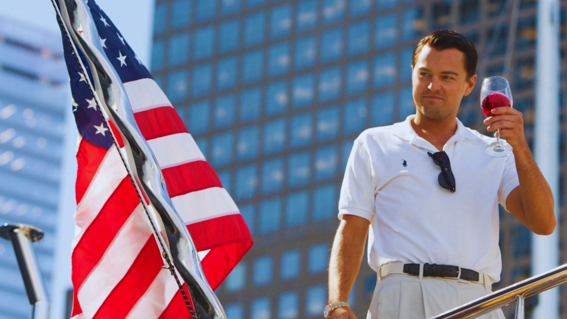 A man in a white polo shirt stands on a boat, holding a glass of red wine, with a confident expression. An American flag waves prominently nearby.