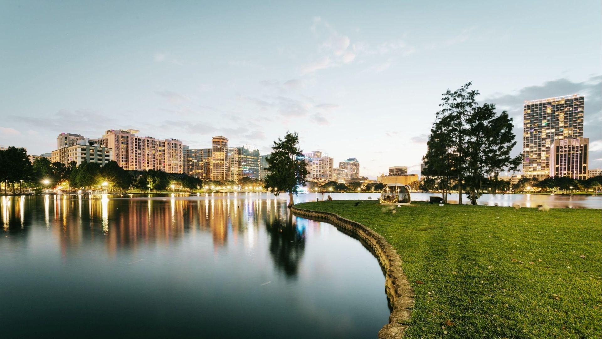 Lakeside city park at dusk with grassy lawn, trees, lake reflections, and illuminated downtown skyline