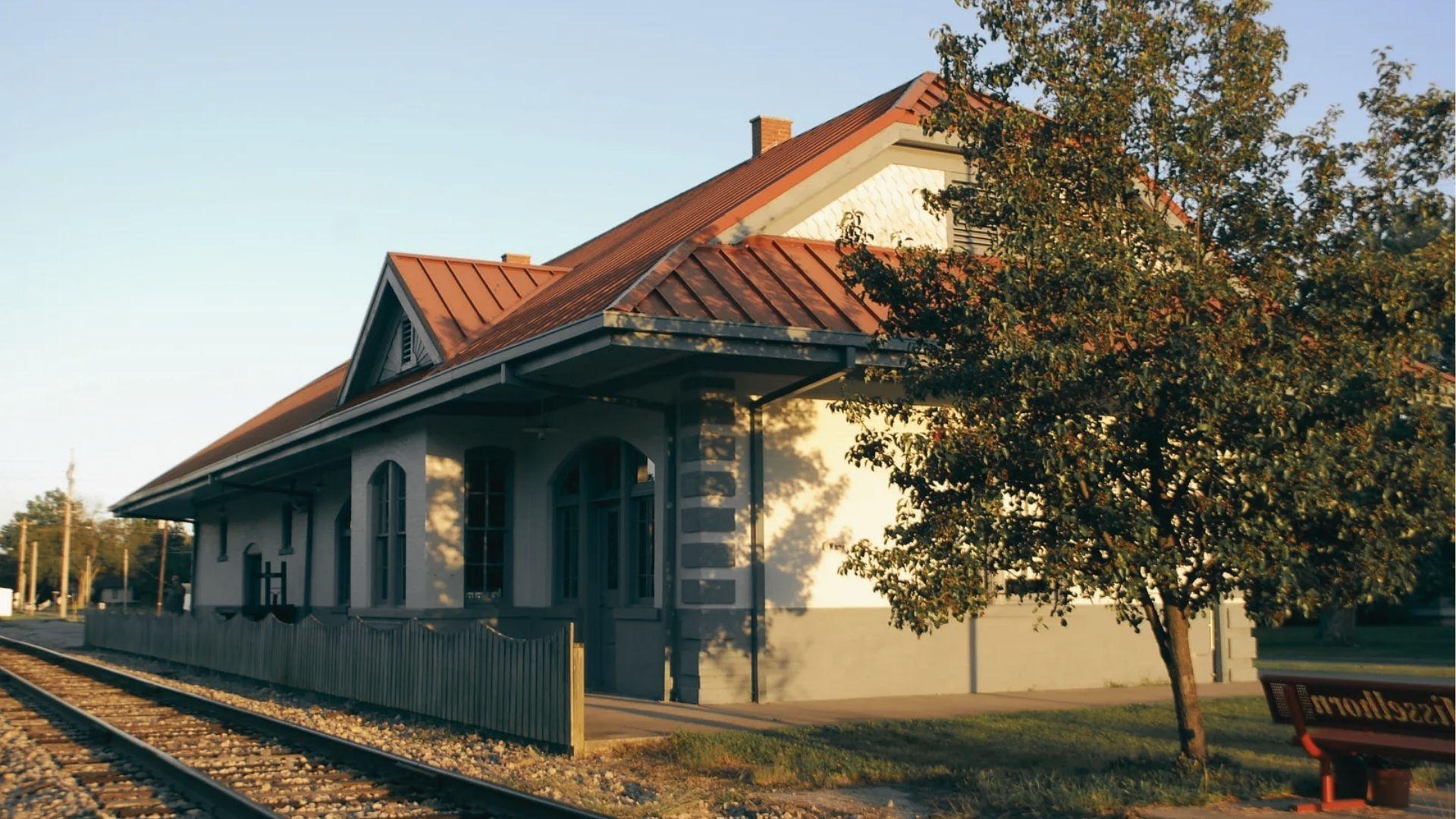 A historic train depot with red metal roof and arched windows beside railroad tracks at golden hour.