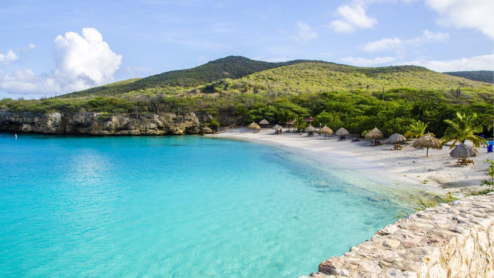 Clear turquoise bay with thatched umbrellas on white sand beach, cliffs, and green hills