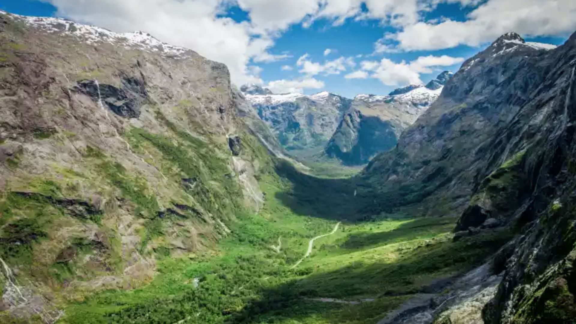 Steep, glacier-carved valley with waterfalls, snow-dusted peaks, and a winding road through lush green forest under a bright blue sky.
