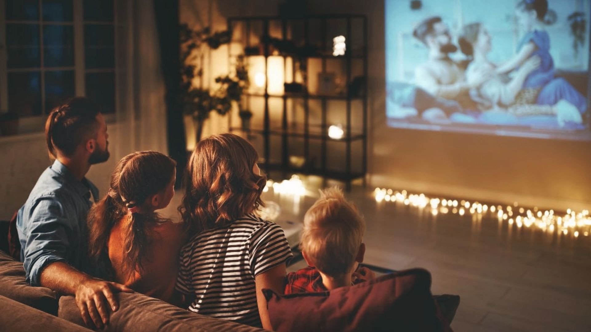 Family of four cuddles on a couch watching a projected movie, surrounded by warm lighting