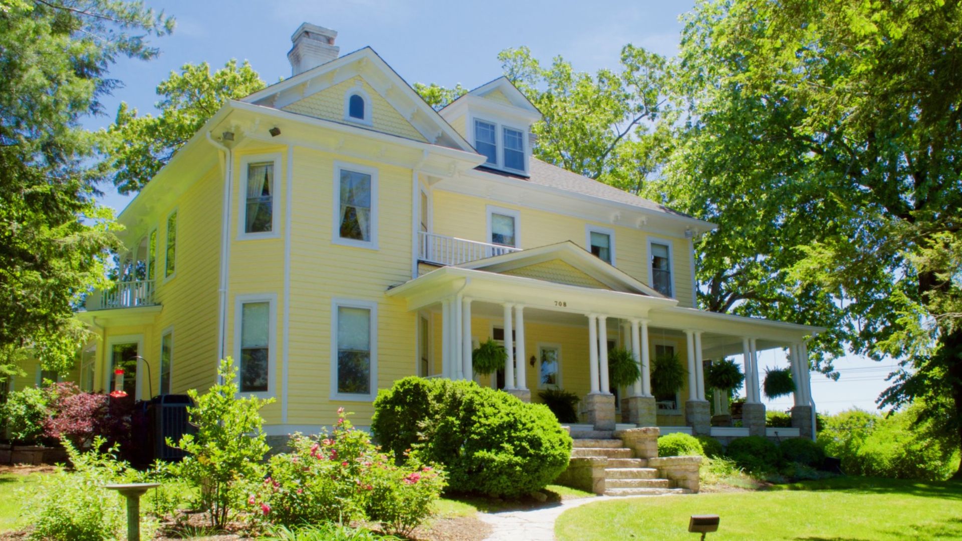 A large yellow Victorian home with white columns and a wraparound porch surrounded by lush gardens.