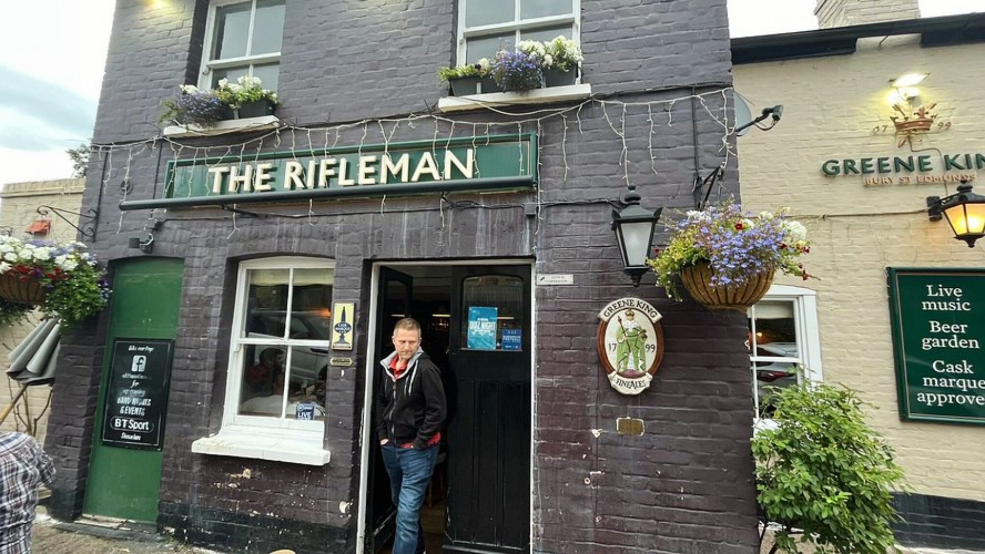 Brick facade of “The Rifleman” pub with hanging flower baskets and Greene King signage.