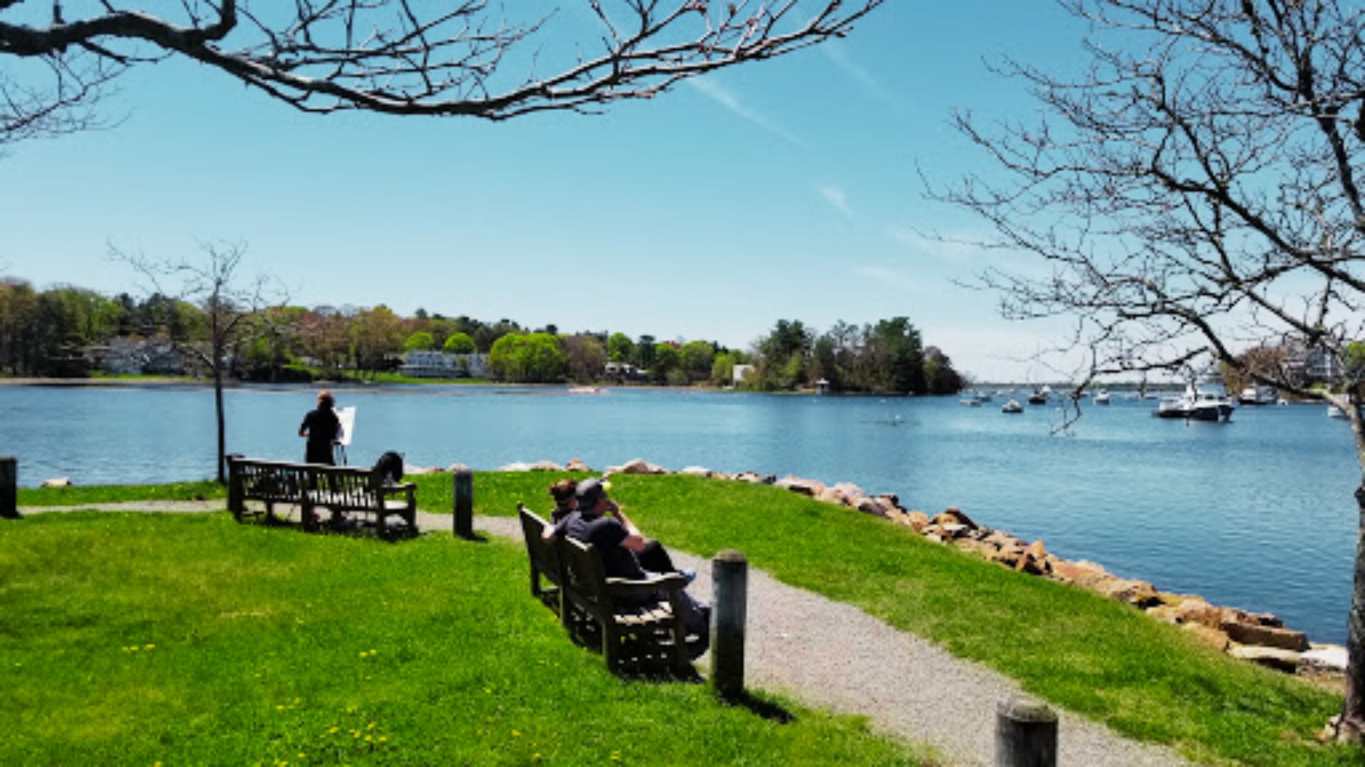 Visitors relax on benches along a grassy waterfront path overlooking Manchester Harbor in Manchester-by-the-Sea.