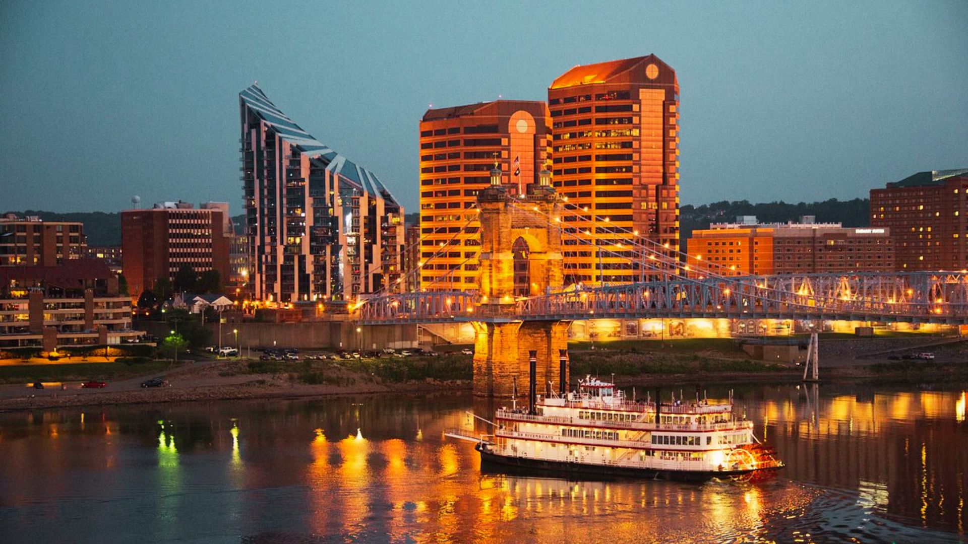 A riverboat cruises the Ohio River at dusk with the Roebling Suspension Bridge and Covington, Kentucky skyline lit up.