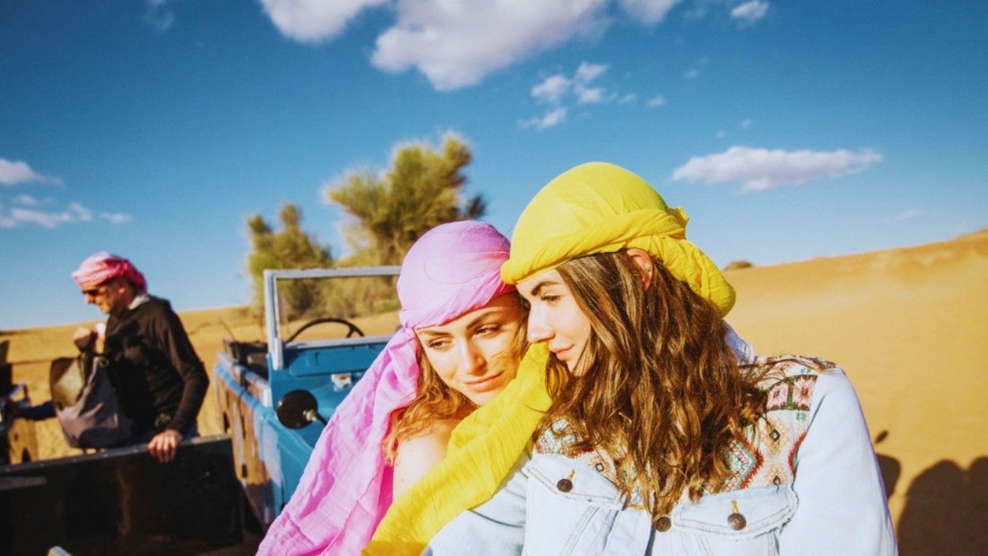 Two women wearing colorful headscarves and hats smile together in a desert, celebrating their best girls trip.