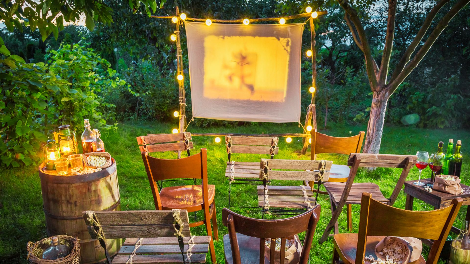String lights illuminate a backyard movie night under a starry sky, with a large projector screen, bean bags, and lawn chairs on the grass.